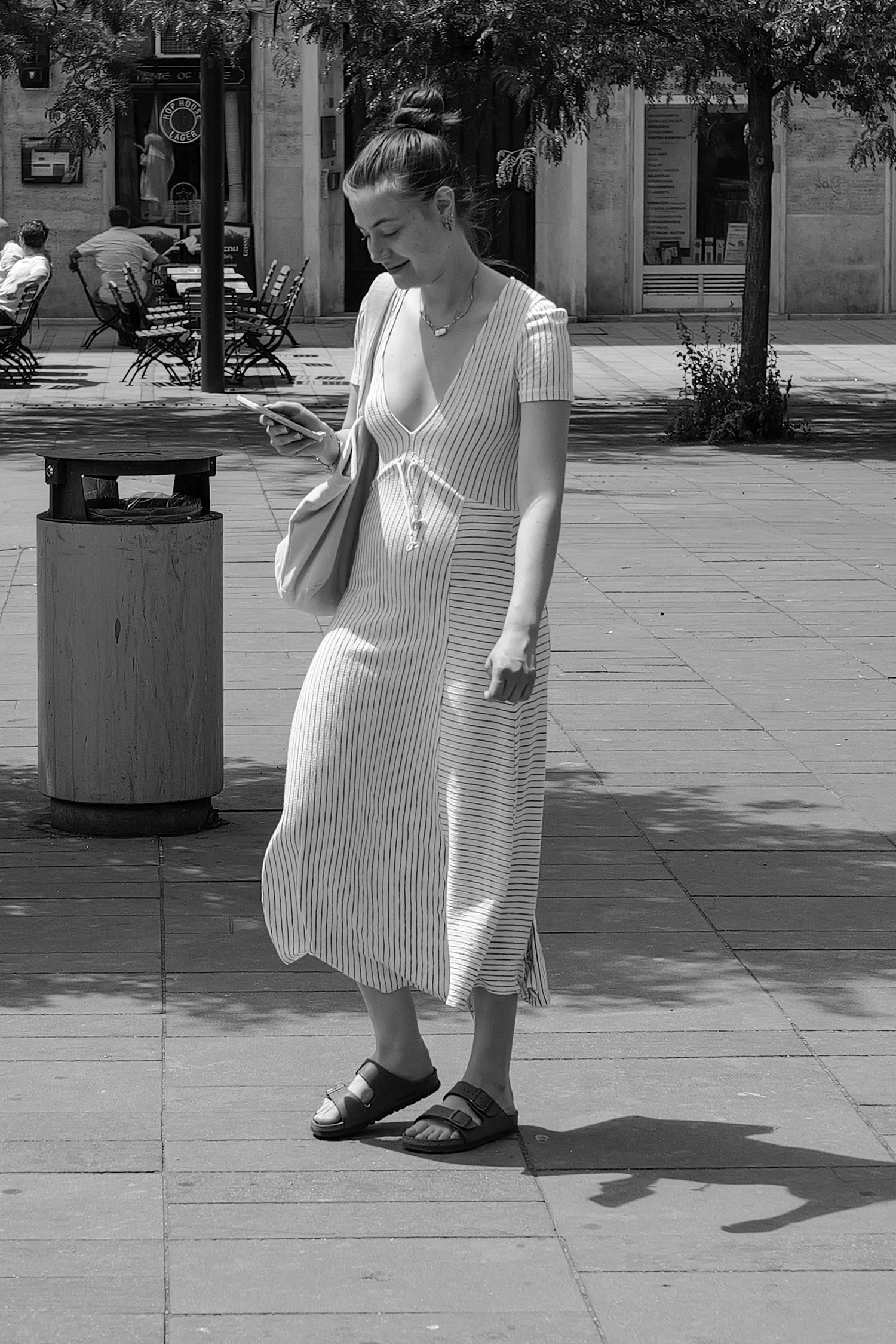 Une jeune femme souriante portant une robe à rayures, des sandales noires et des cheveux attachés, marche dans une rue ensoleillée, tenant un téléphone dans une main.