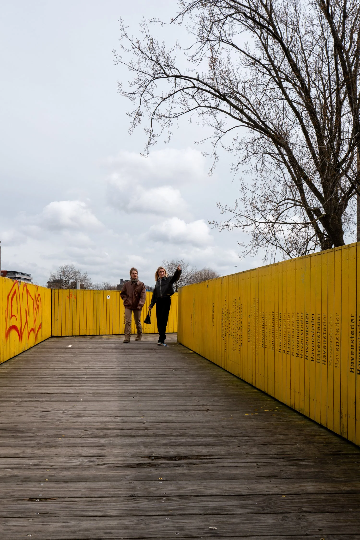 Deux femmes marchent sur un pont en bois bordé de murs jaunes avec graffiti et texte en anglais noir, arbres dénudés et ciel nuageux en arrière-plan.
