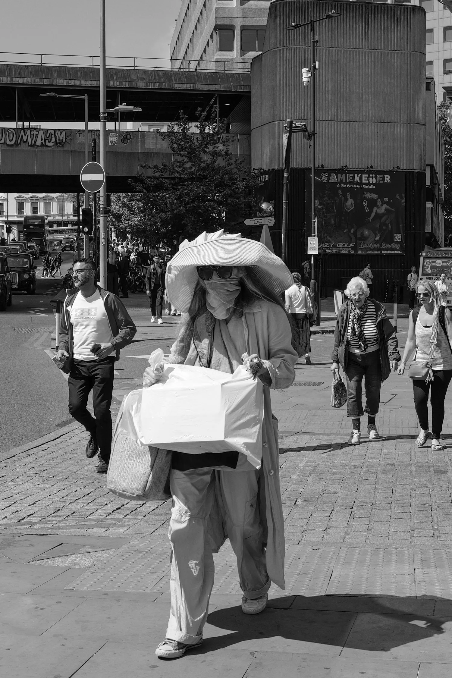 Une femme portant un grand chapeau, des lunettes de soleil et un masque, qui pousse une poussette dans une rue animée d'une ville