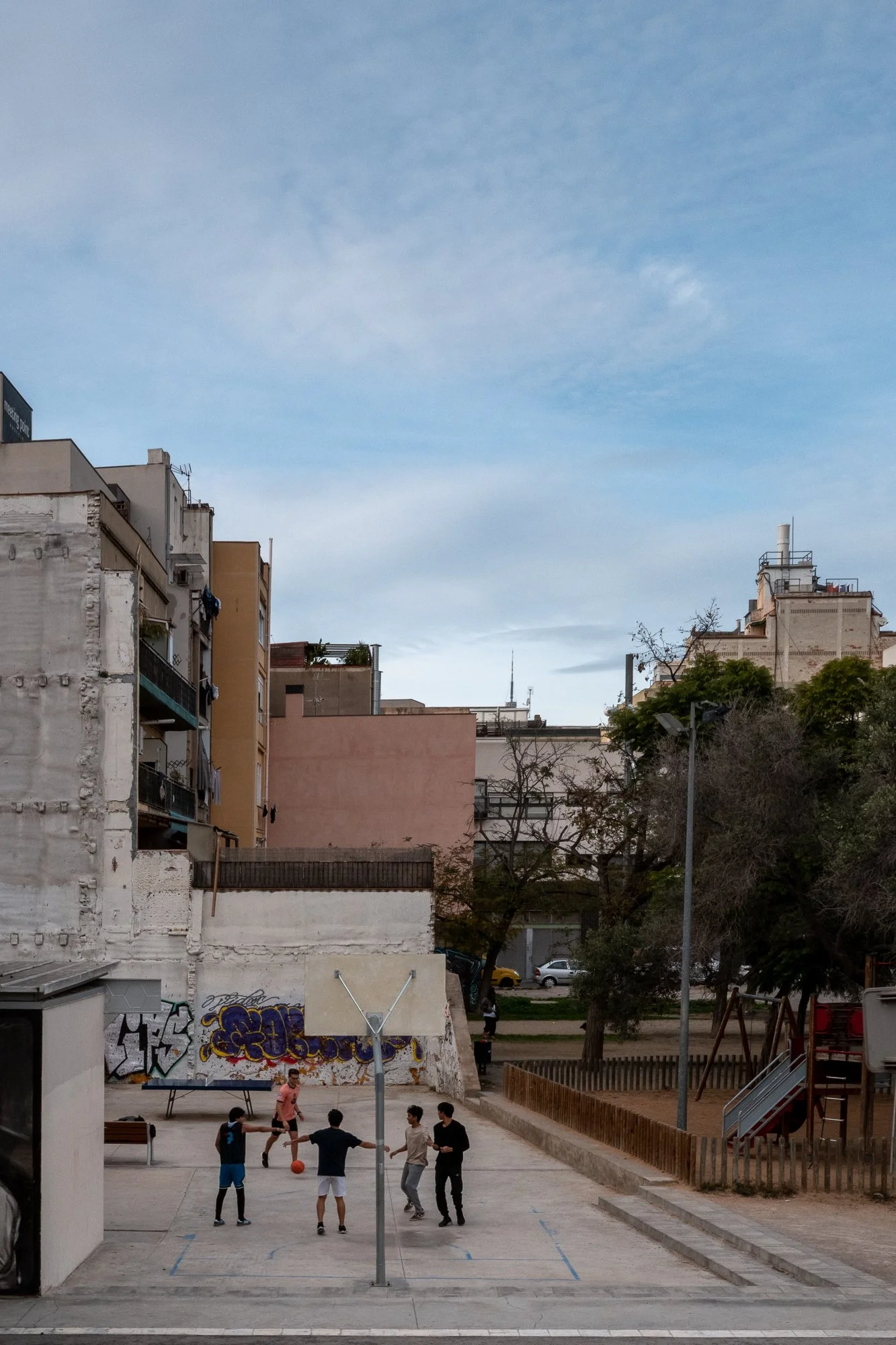Des enfants jouent au basketball dans une cour d'école urbaine avec des graffitis sur un mur, des arbres, et des bâtiments résidentiels en arrière-plan, sous un ciel partiellement nuageux.