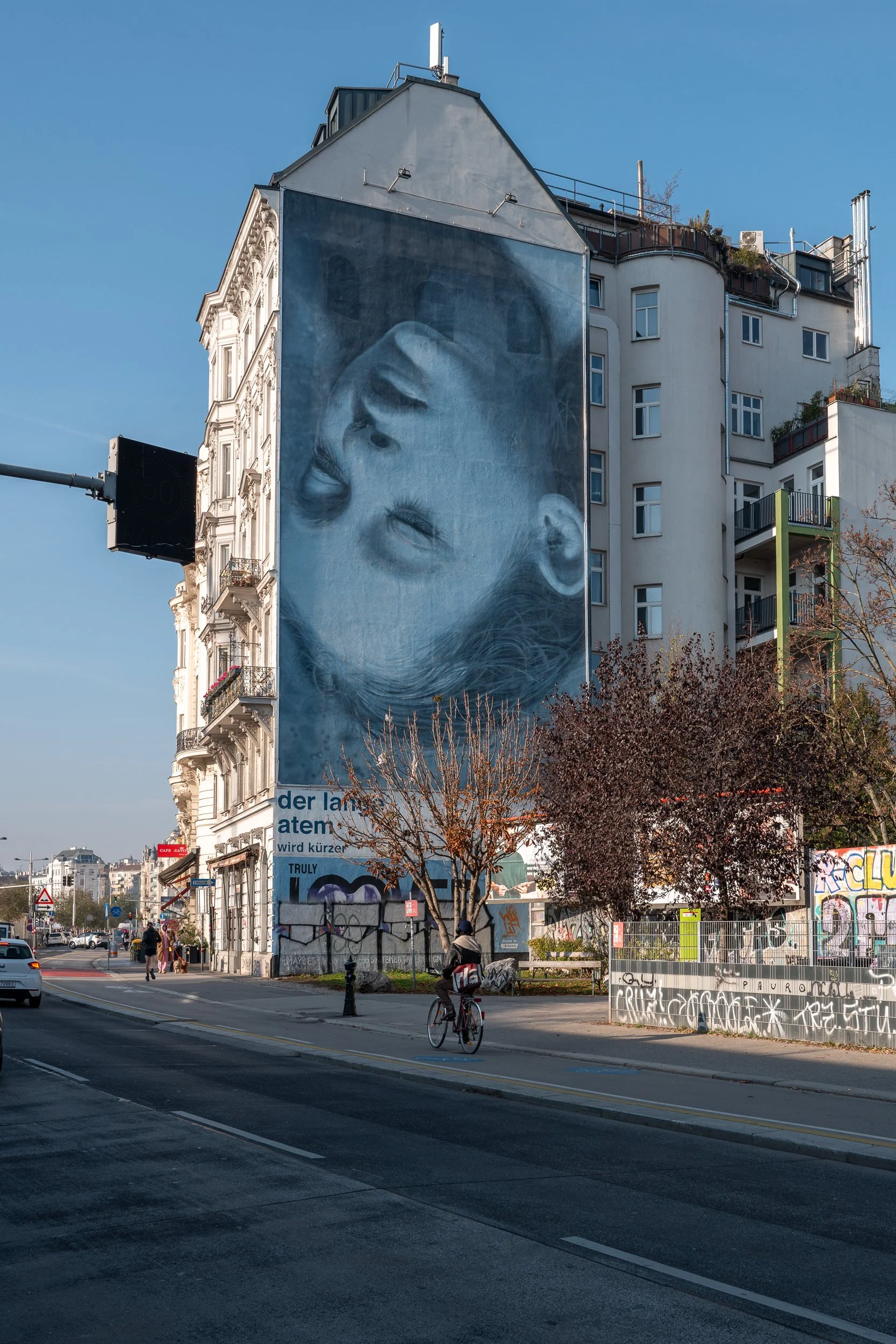 Mur de rue avec une grande affiche artistique représentant le visage d’une enfant en noir et blanc, dans une zone urbaine avec des arbres, des bâtiments modernes et un cycliste.