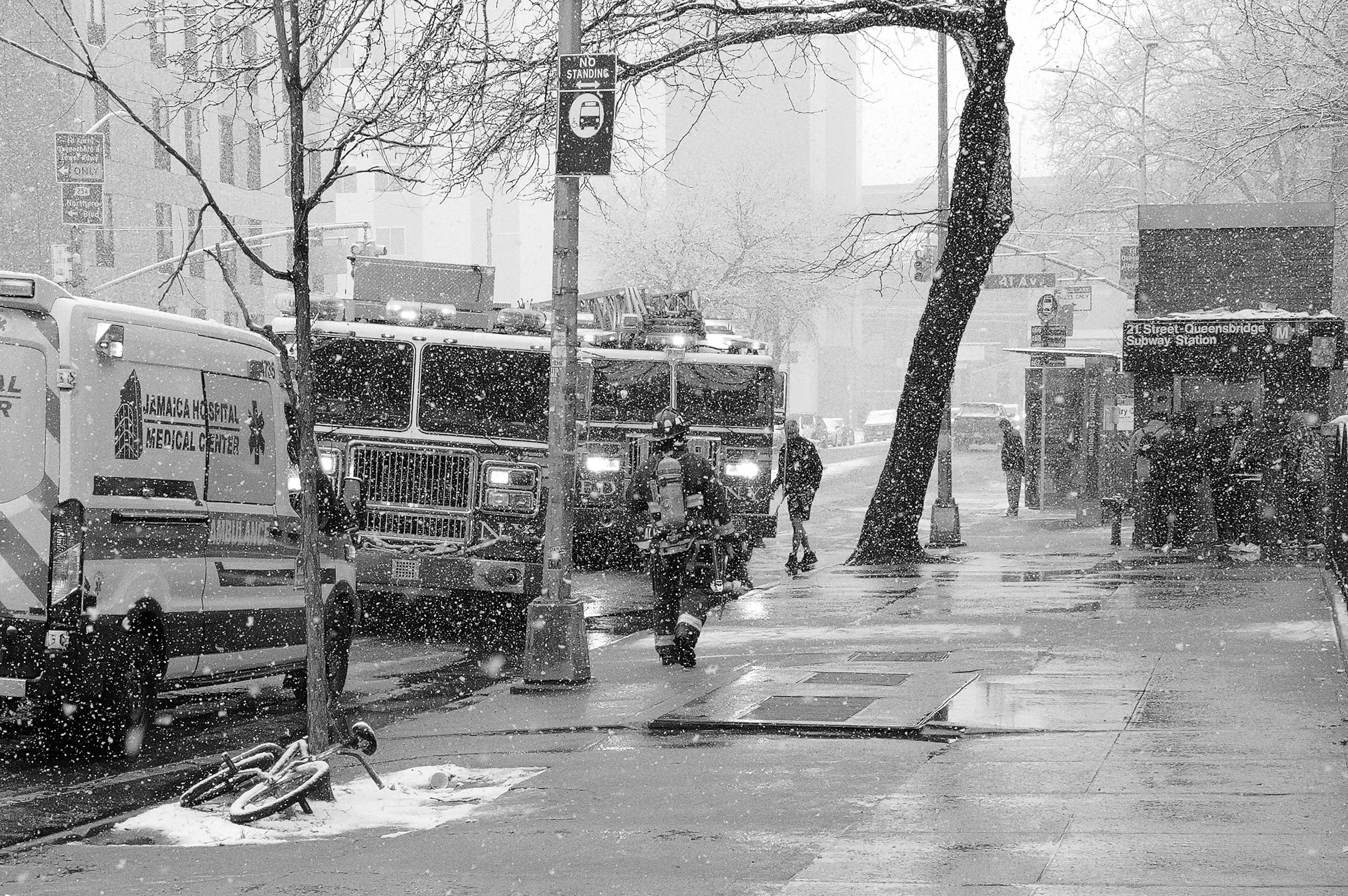 Ambulance et camion de pompiers en service lors d'une tempête de neige dans la rue, avec des passants, un arbre et un abri-bus.