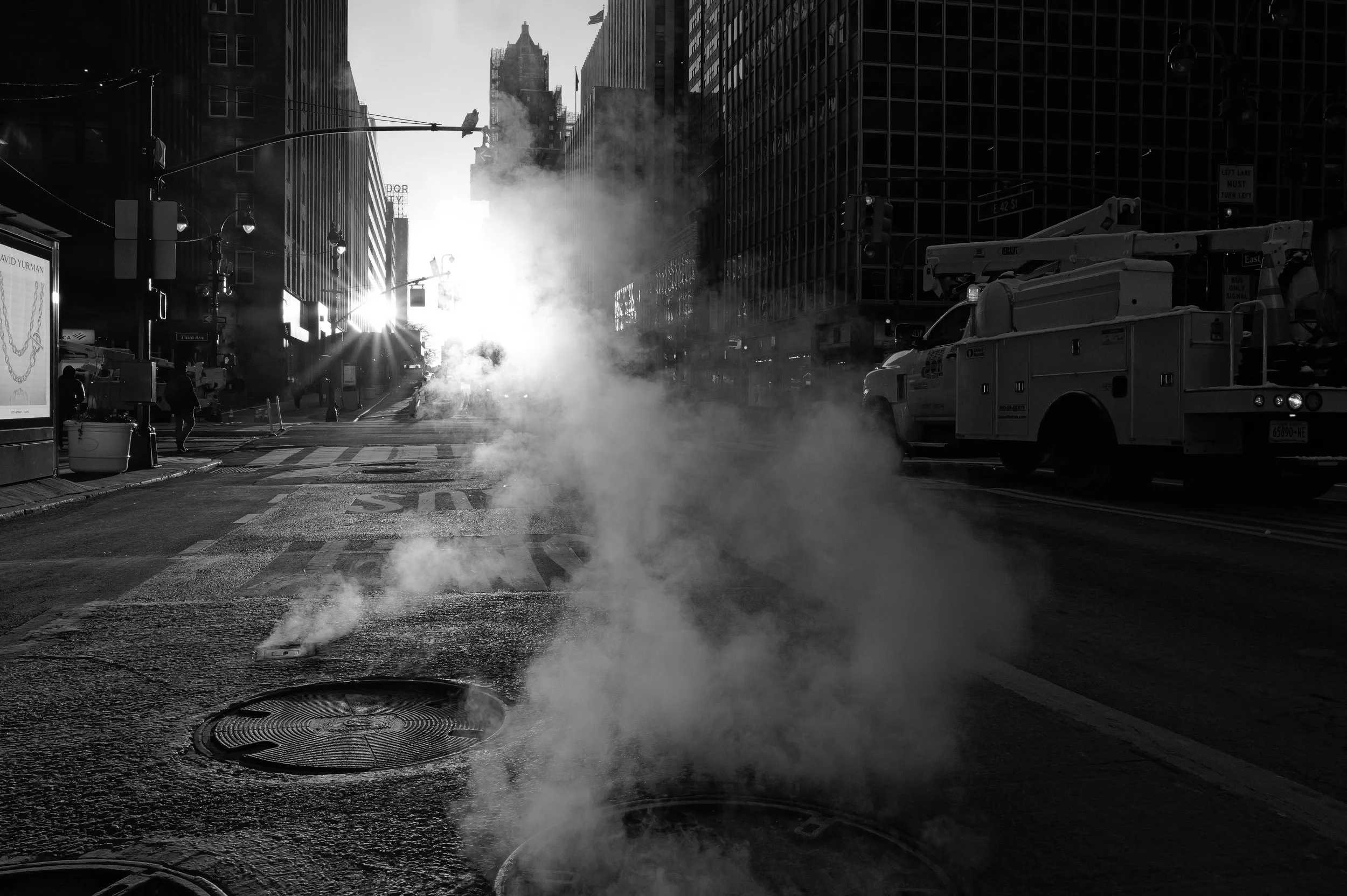 Une rue urbaine en noir et blanc avec de la vapeur qui s'échappe de bouches d'aération, des bâtiments modernes et une lumière intense du soleil à l'horizon
