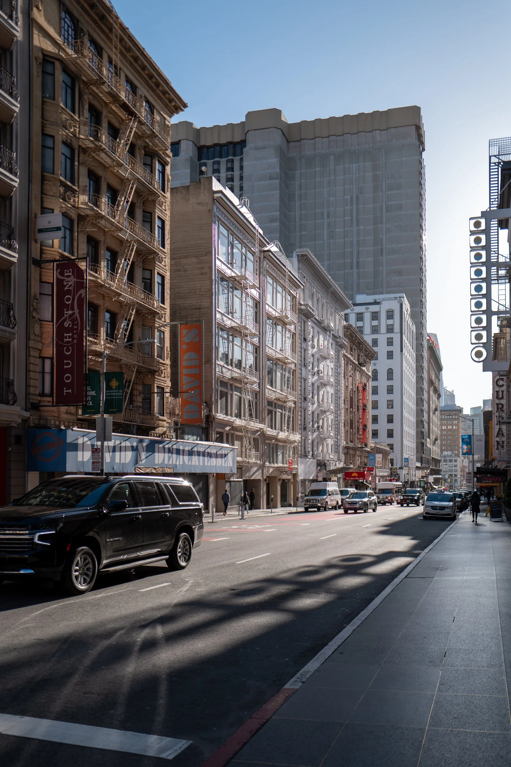Une rue urbaine avec des immeubles, des voitures stationnées et des passants, sous un ciel clair.