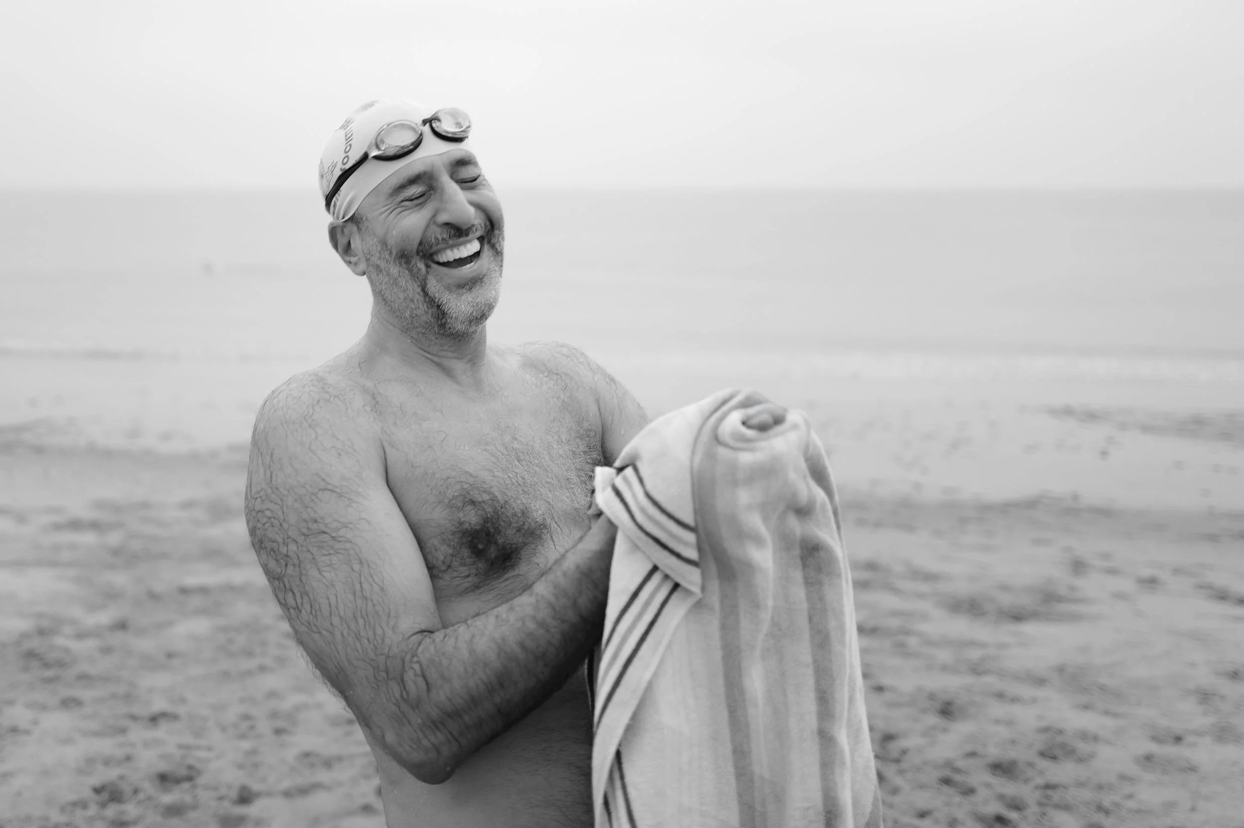Un homme souriant, sans chemise, portant un bonnet de bain avec des lunettes de natation dessus, tenant une serviette sur la plage.