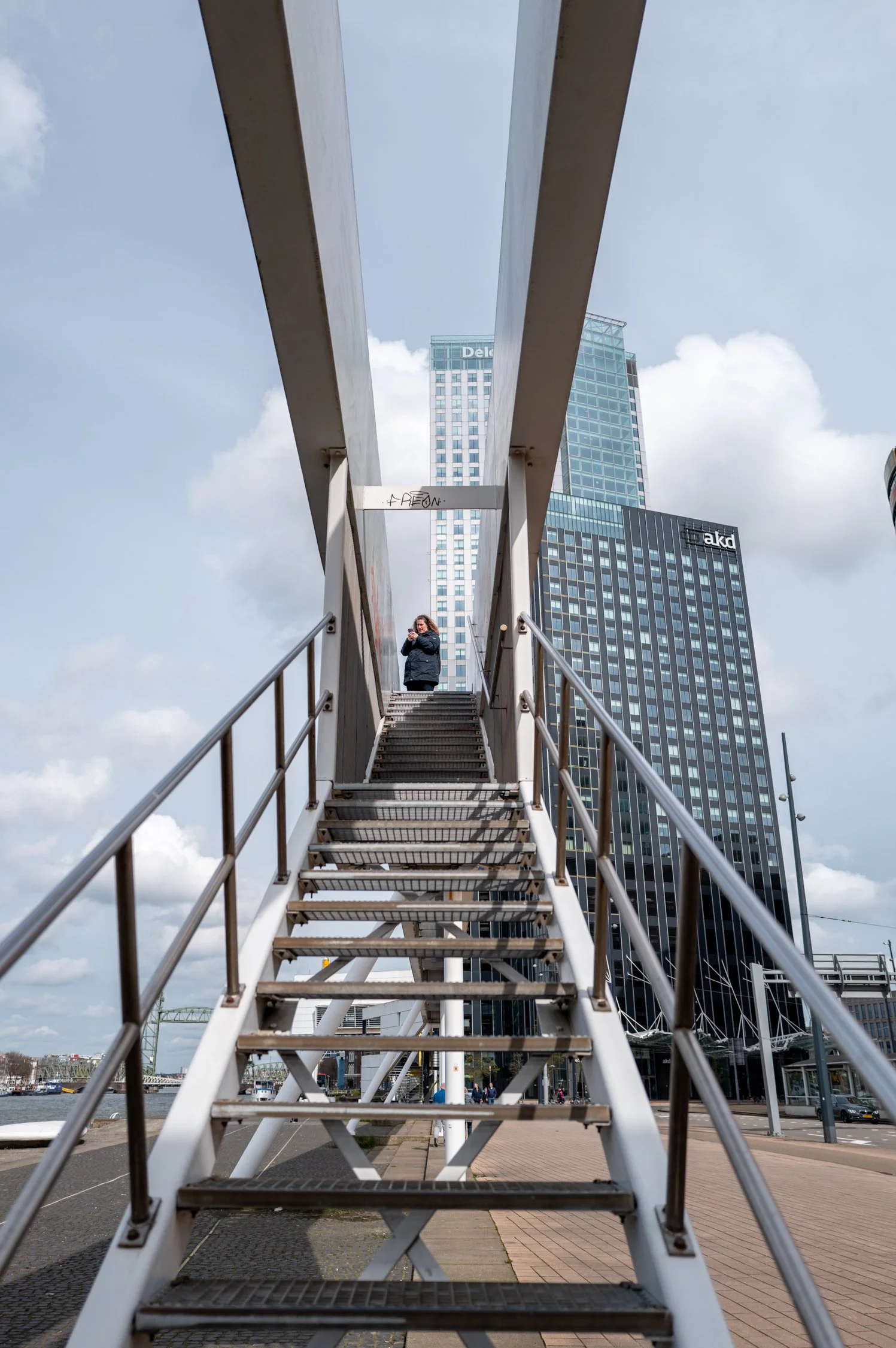 Une passerelle piétonne en acier vue de dessous, avec un homme debout en haut, devant des gratte-ciel modernes sous un ciel nuageux