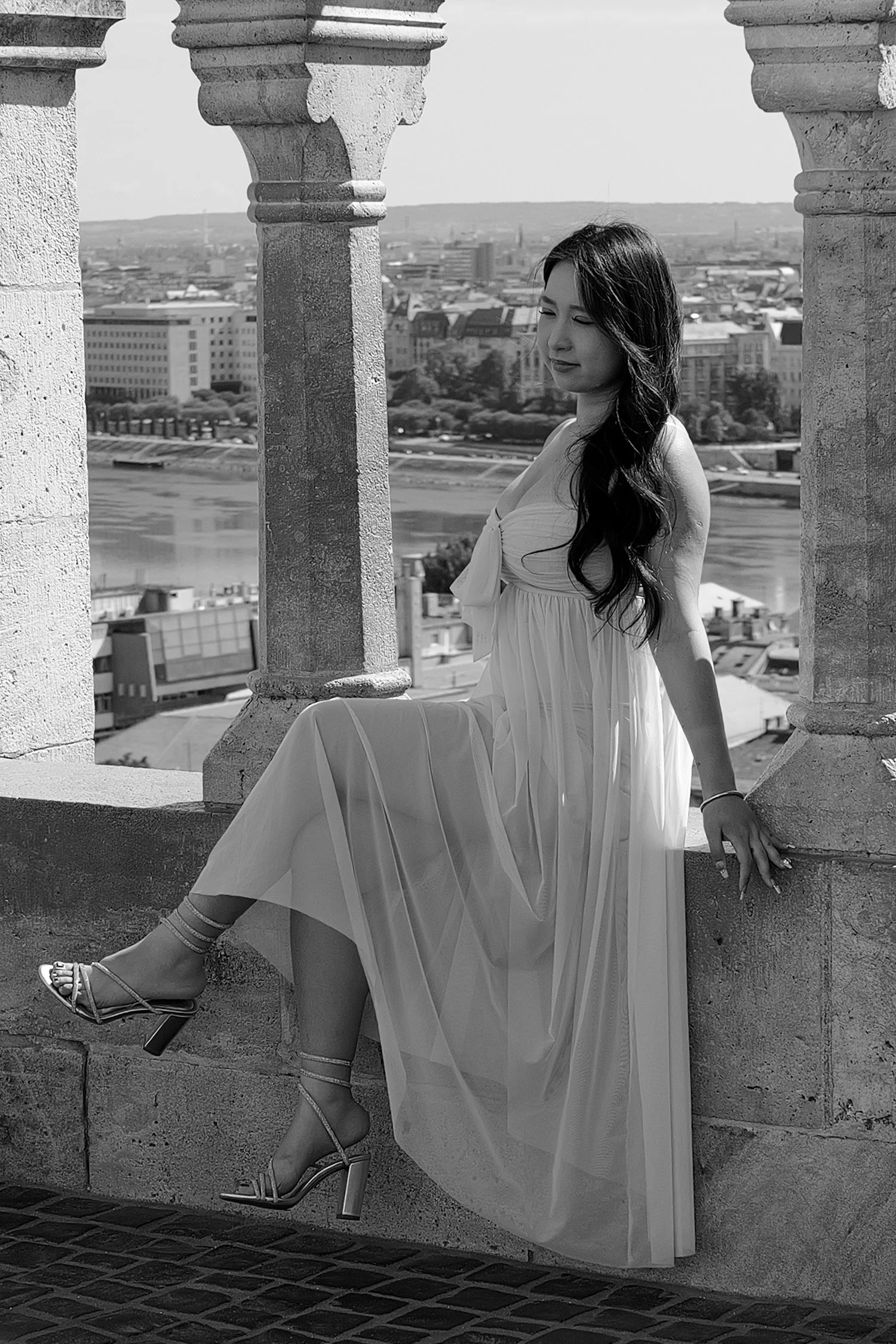 Une femme en robe longue assise sur une balustrade en pierre, avec une ville en arrière-plan, photographiée en noir et blanc.