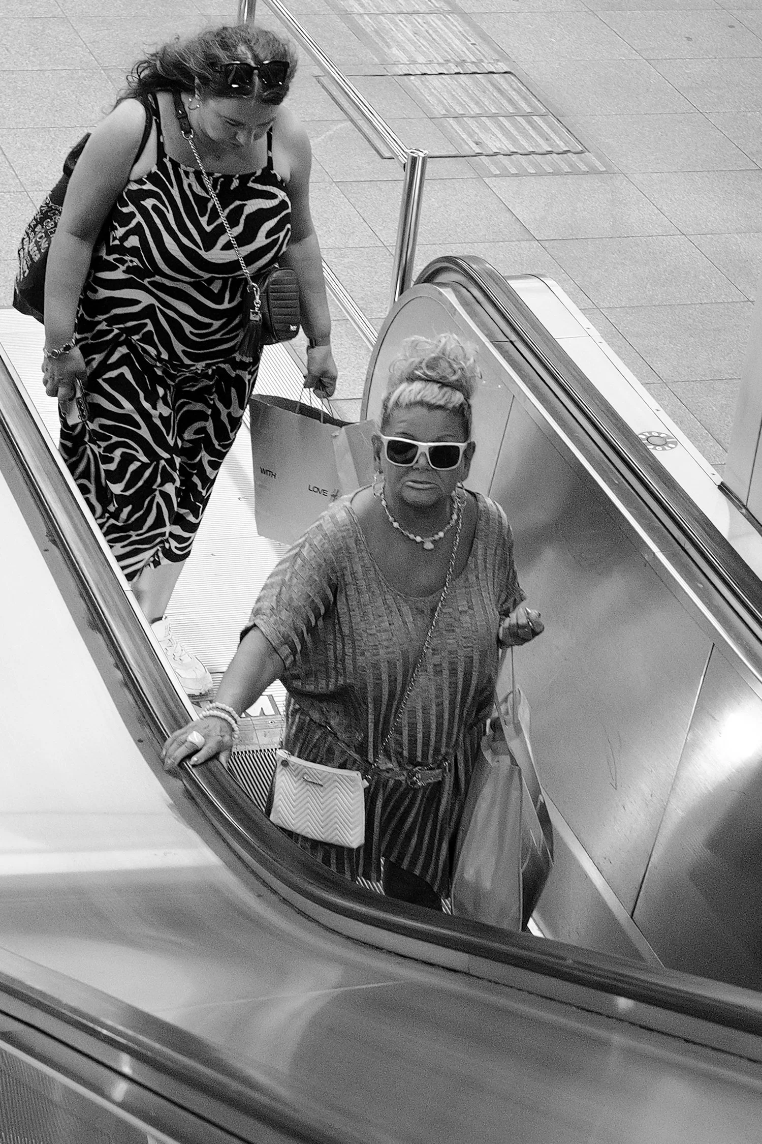 Deux femmes avec des vêtements à motifs et des accessoires voyants, visibles dans un miroir d'ascenseur, l'une avec des lunettes foncées et une coiffure en chignon, l'autre portant des lunettes de soleil blanches et une coiffure en chignon aussi.