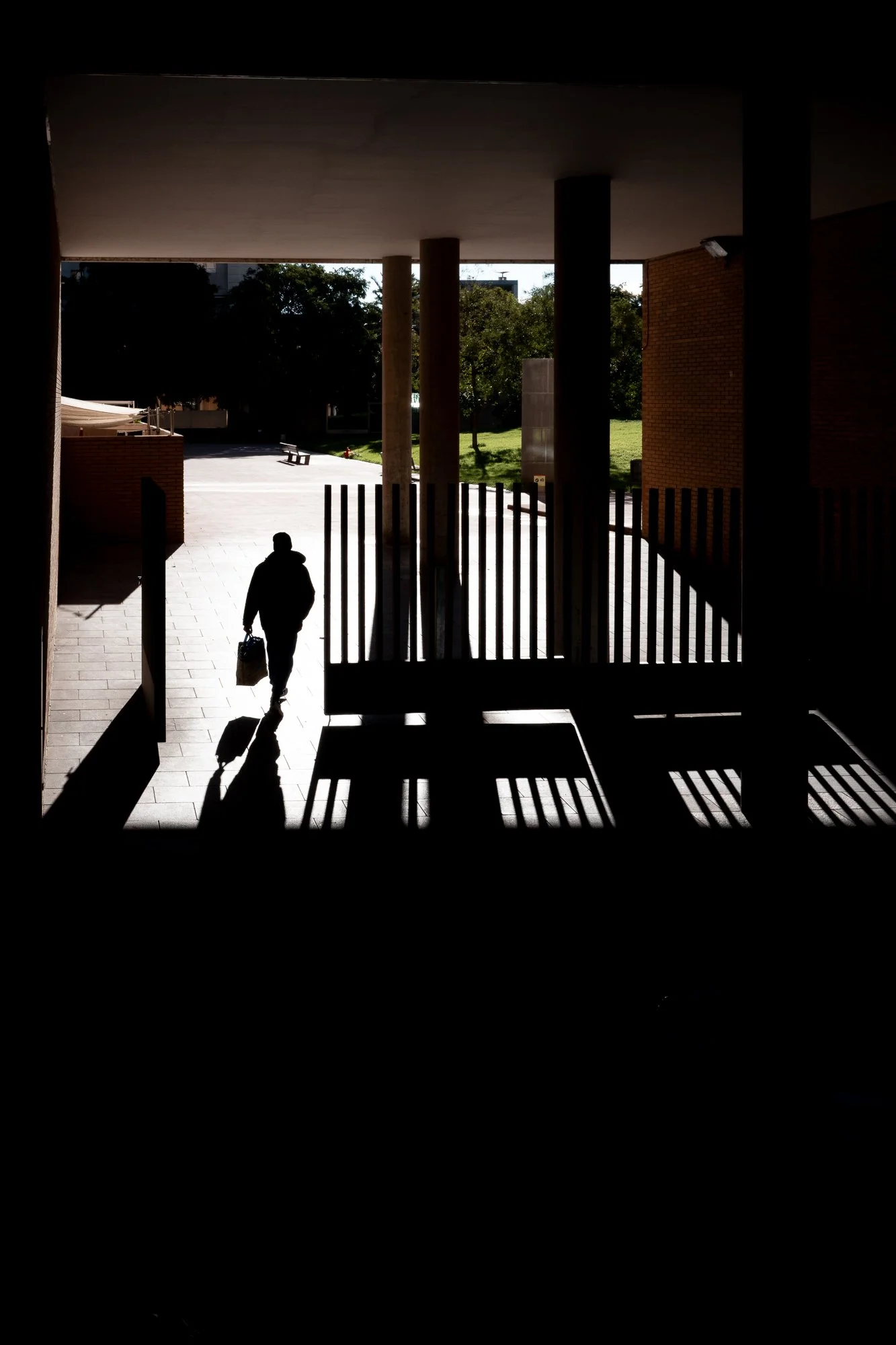 Silhouette d'une personne marchant avec un sac en main à l'extérieur, vue depuis l'intérieur d'un bâtiment vers un espace extérieur avec des arbres et un chemin ensoleillé.