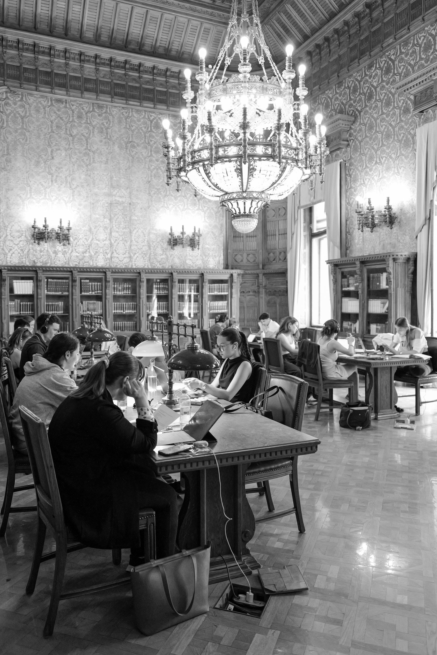 Une bibliothèque ancienne luxueuse avec un plafond en bois, un grand chandelier, des étagères remplies de livres, et des étudiants travaillant à des tables.