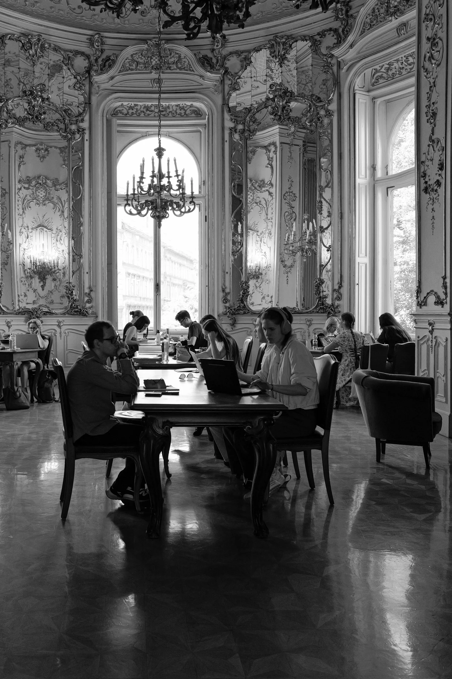 Une salle élégante avec des murs ornés de miroirs et de moulures baroques, un plafond décoré de moulures, un grand chandelier en cristal, et plusieurs personnes assises à des tables, travaillant ou étudiant, éclairées par la lumière naturelle provena
