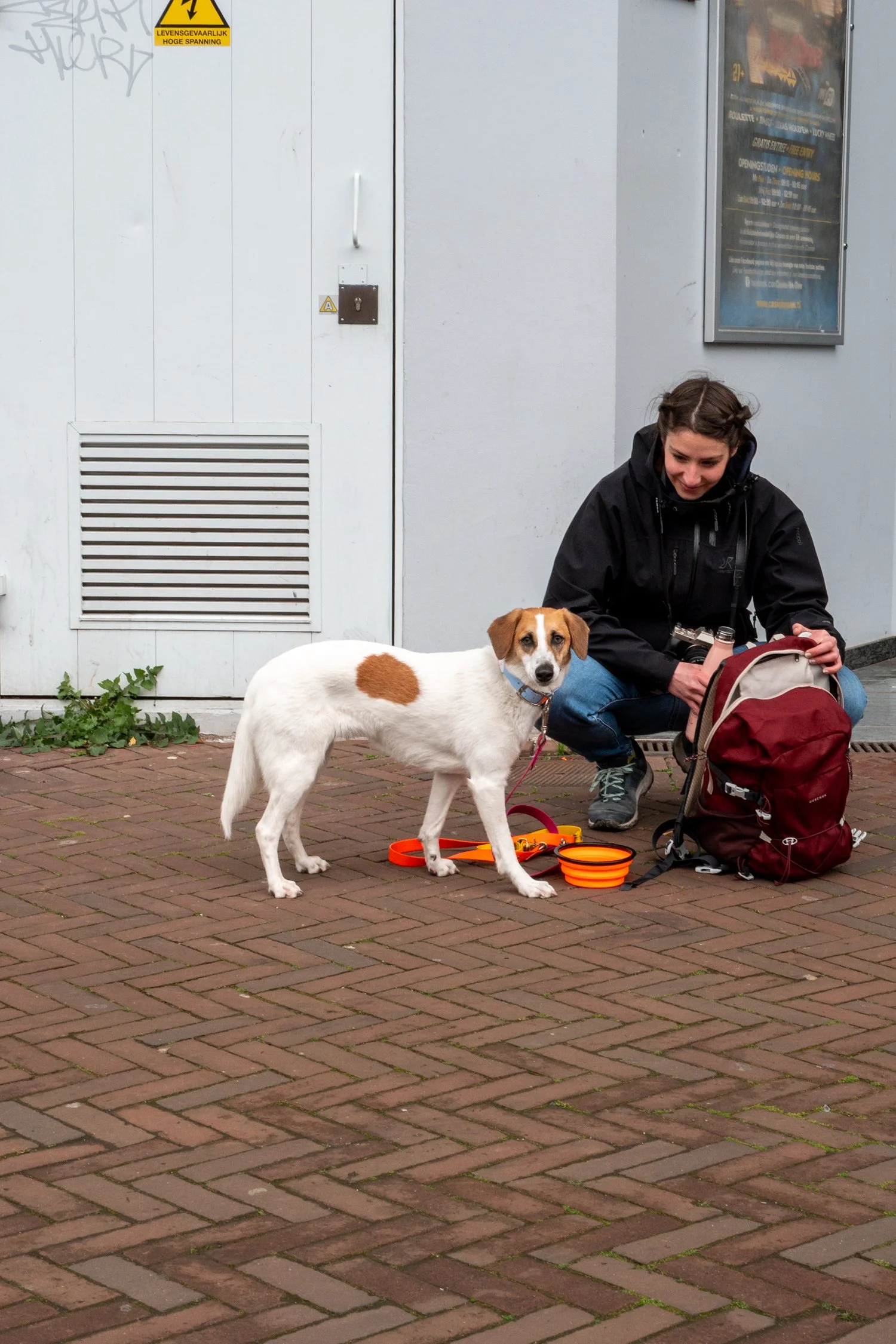 Une femme souriante, assise sur le trottoir, avec un chien blanc et marron. La femme porte un manteau noir et une montre, et elle sort un appareil photo d'un sac à dos rouge. Le chien porte un collier bleu et se tient près d'une gamelle orange. Il y 