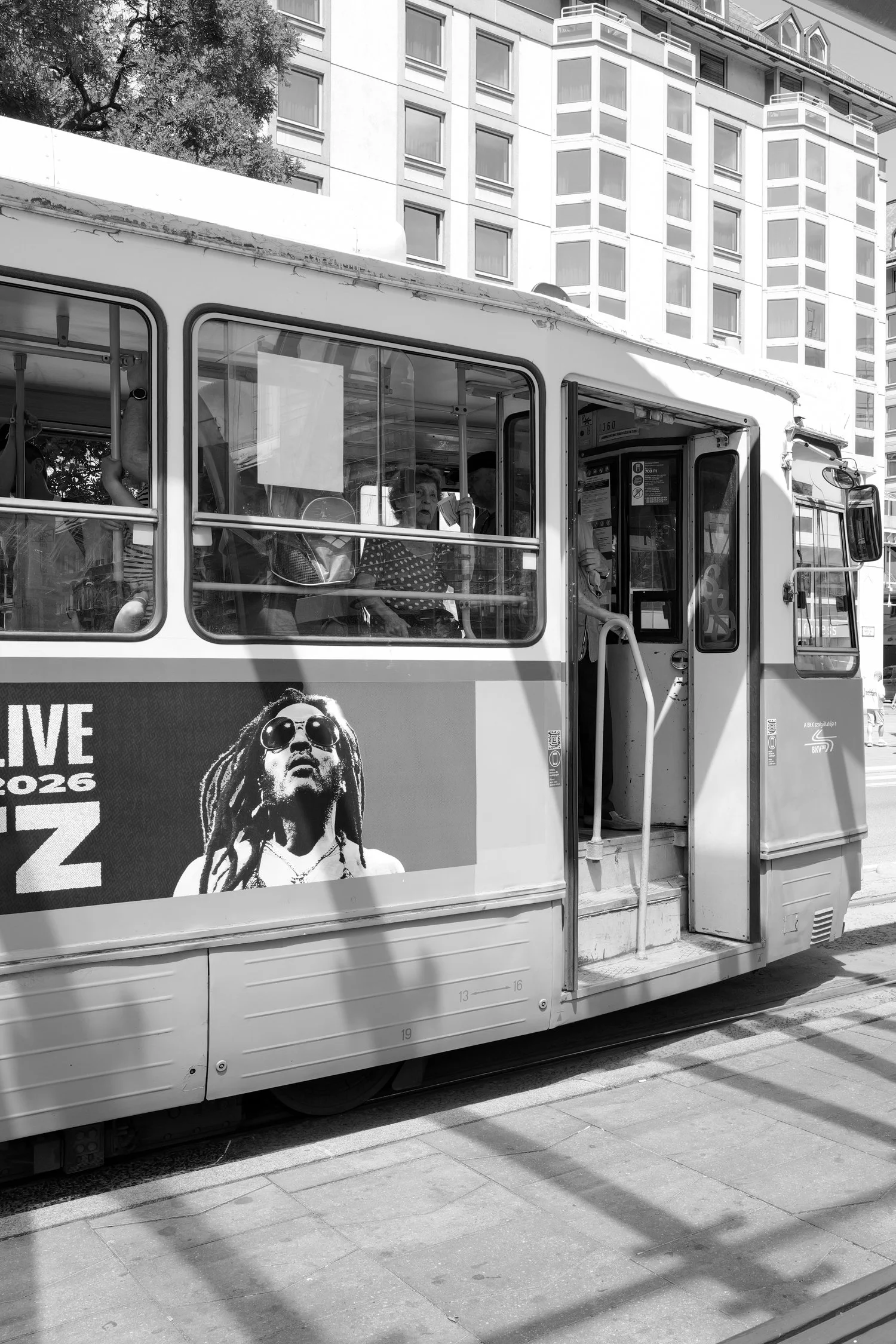Un tramway en noir et blanc avec une affiche promotionnelle d'un concert de musique avec une image d'un homme avec des dreadlocks et des lunettes de soleil, situé dans une zone urbaine avec des immeubles résidentiels en arrière-plan.