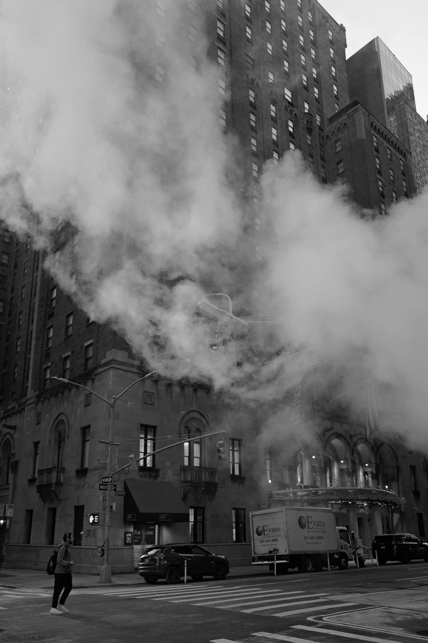 Une rue urbaine avec un bâtiment en feu, de la fumée s'échappant, des voitures stationnées et un piéton portant un sac à dos.