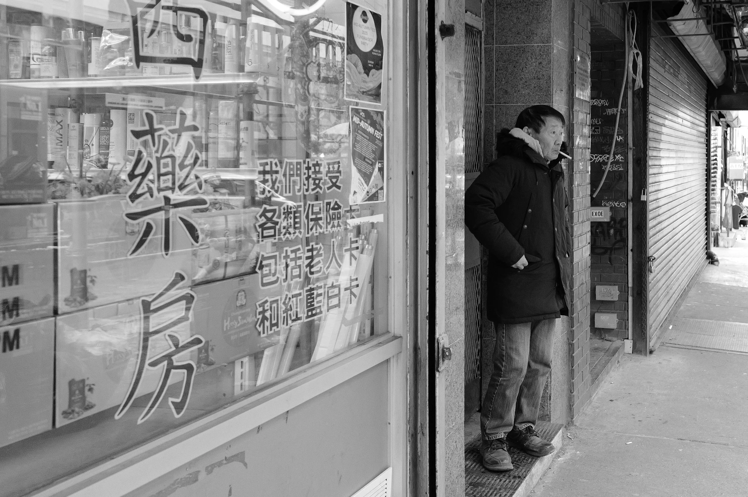 Un homme portant un manteau noir se tient debout sur le trottoir à côté d'une vitrine d'une boutique. La boutique a des affiches en chinois dans la vitre. La scène est en noir et blanc.