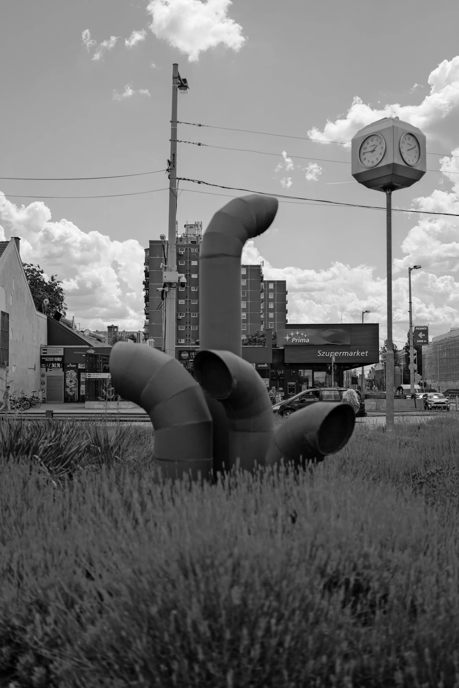 Une photographie en noir et blanc montrant un marteau dans l'herbe, avec une horloge à côté, un bâtiment en arrière-plan et un ciel nuageux.