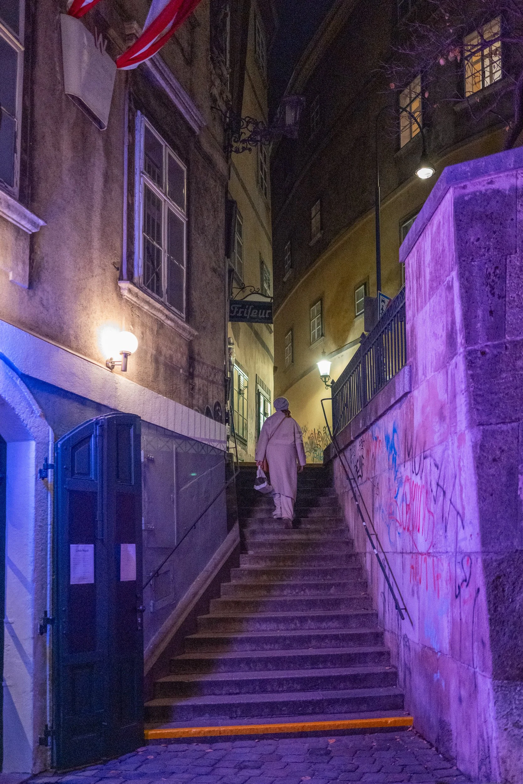 Personne marchant dans un escalier urbain éclairé par des lampes de nuit, murs en pierre et graffiti, nuit avec un éclairage violet et jaune.