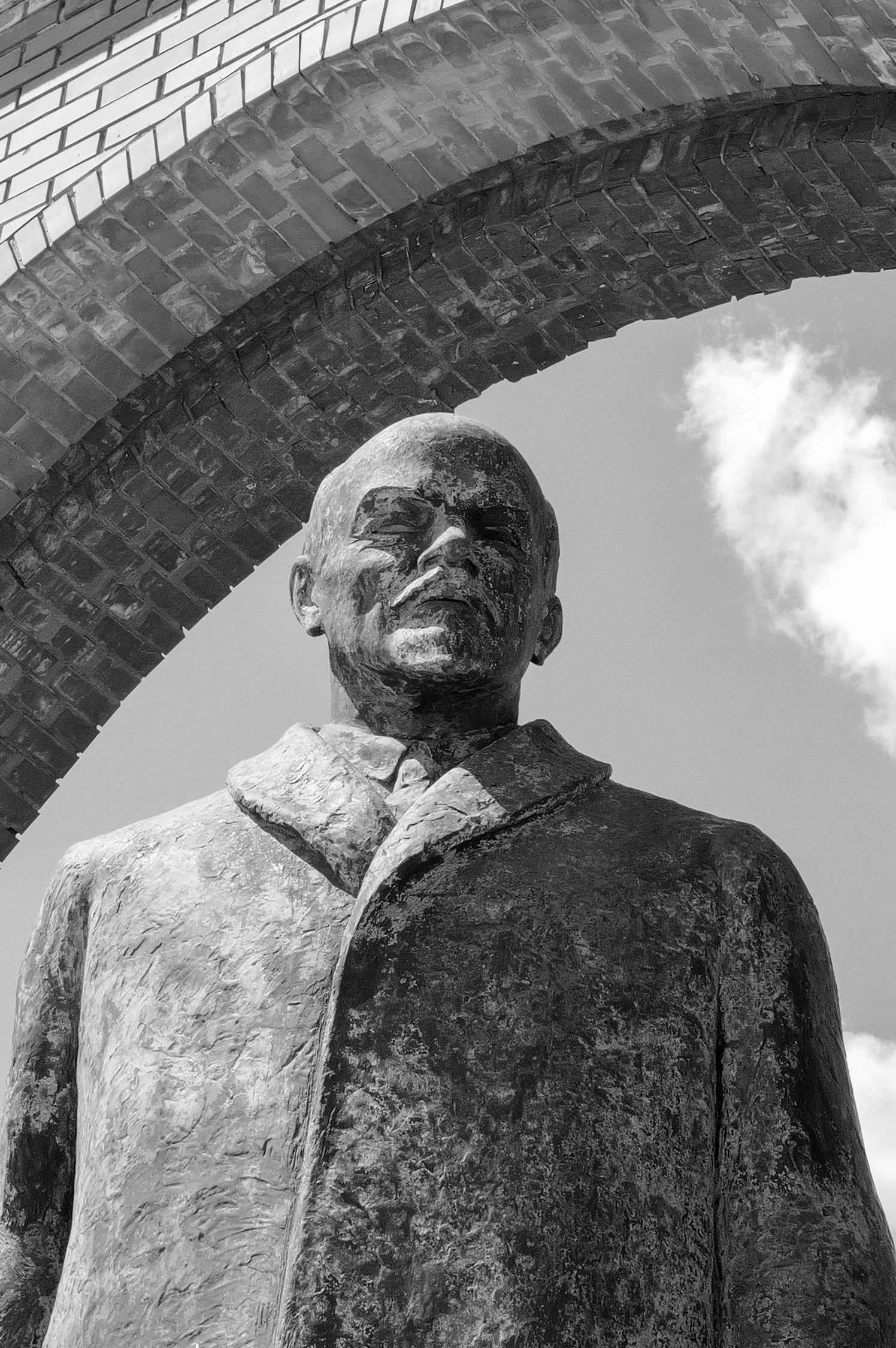 Statue en bronze d'un homme portant un costume avec un nœud de cravate, en face d'une arche en brique, avec un ciel nuageux en arrière-plan.