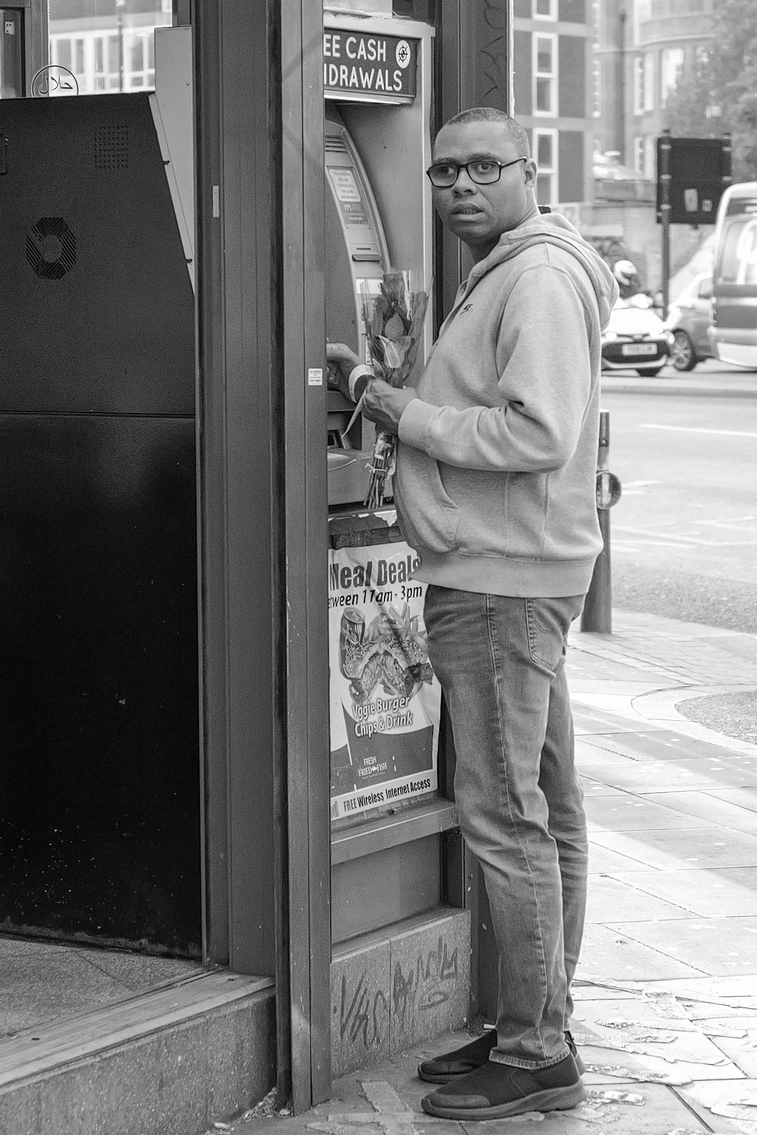 Un homme porte un sweat à capuche et des jeans, en train d'utiliser un distributeur automatique de billets à l'extérieur d'une banque ou d'un guichet automatique. Il tient un bouquet de fleurs dans la main gauche.