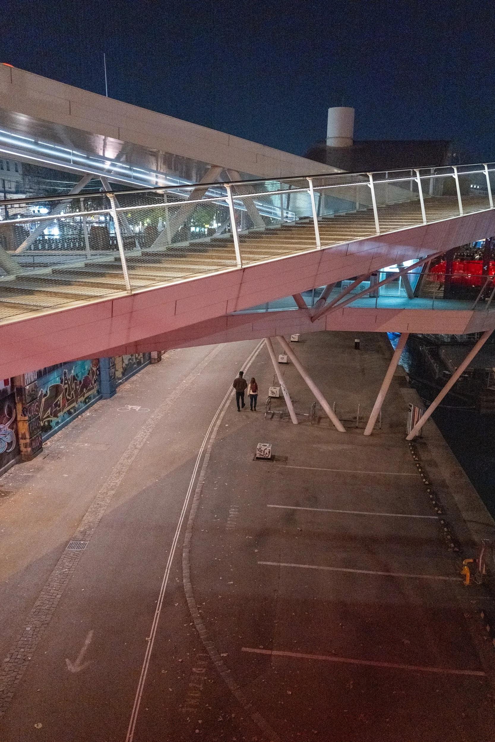 Ville de nuit avec pont piétonnier en spirale, deux personnes marchant, parking vide, graffiti sur les murs, ciel nocturne.