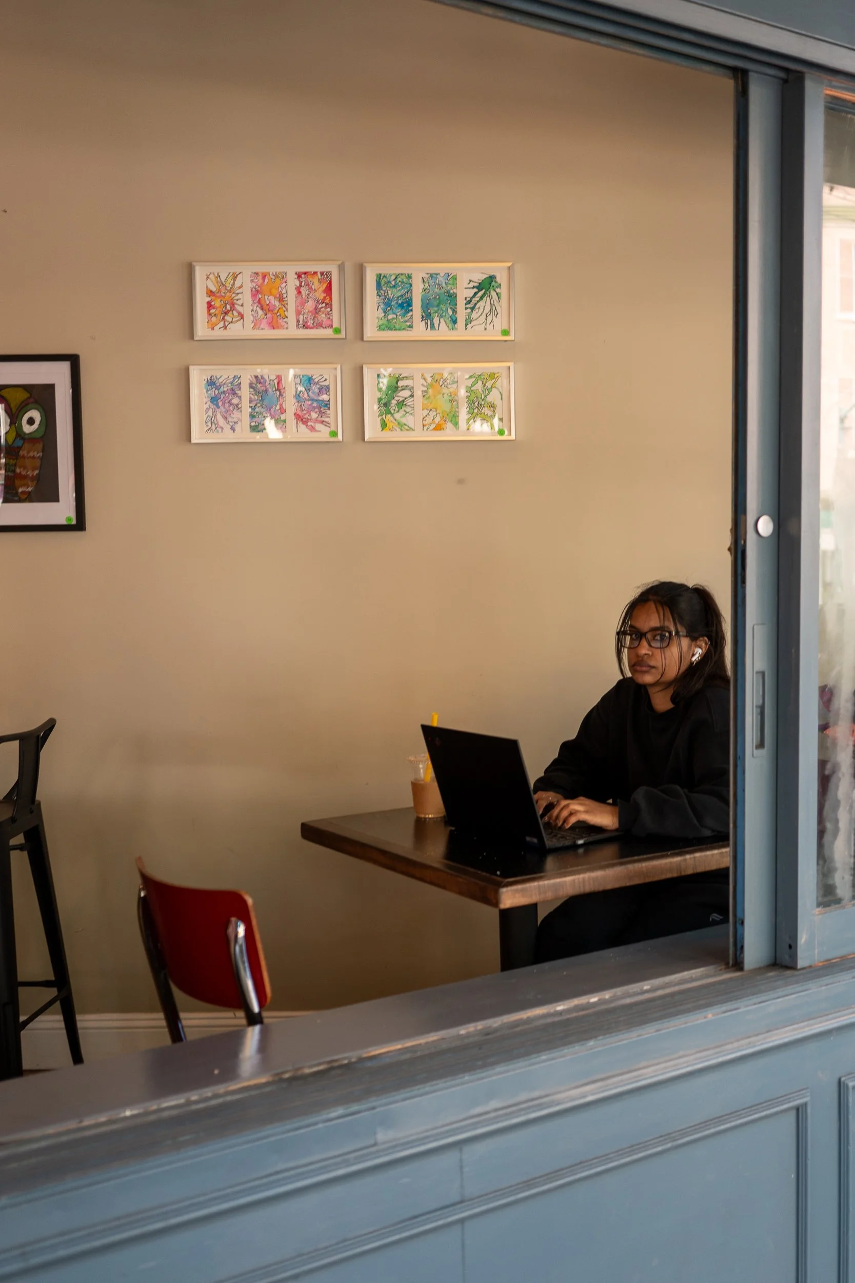 Une femme assise à un bureau dans un café, travaillant sur un ordinateur portable, avec des tableaux colorés accrochant au mur derrière elle.