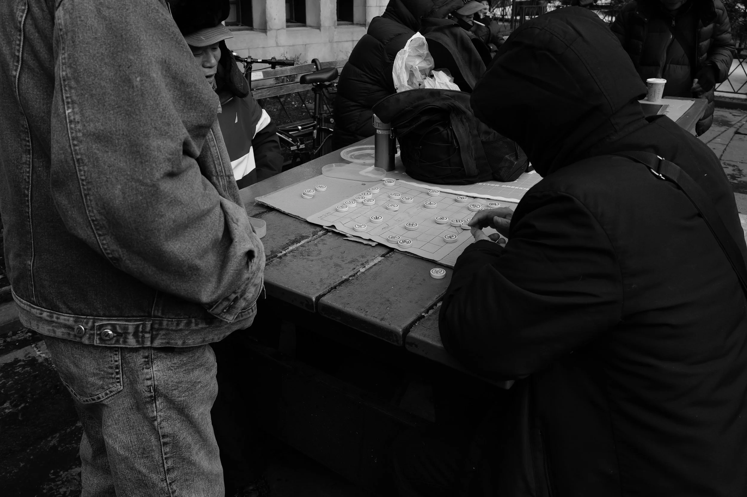 Groupe de personnes jouant aux dames sur une table en extérieur, en noir et blanc.