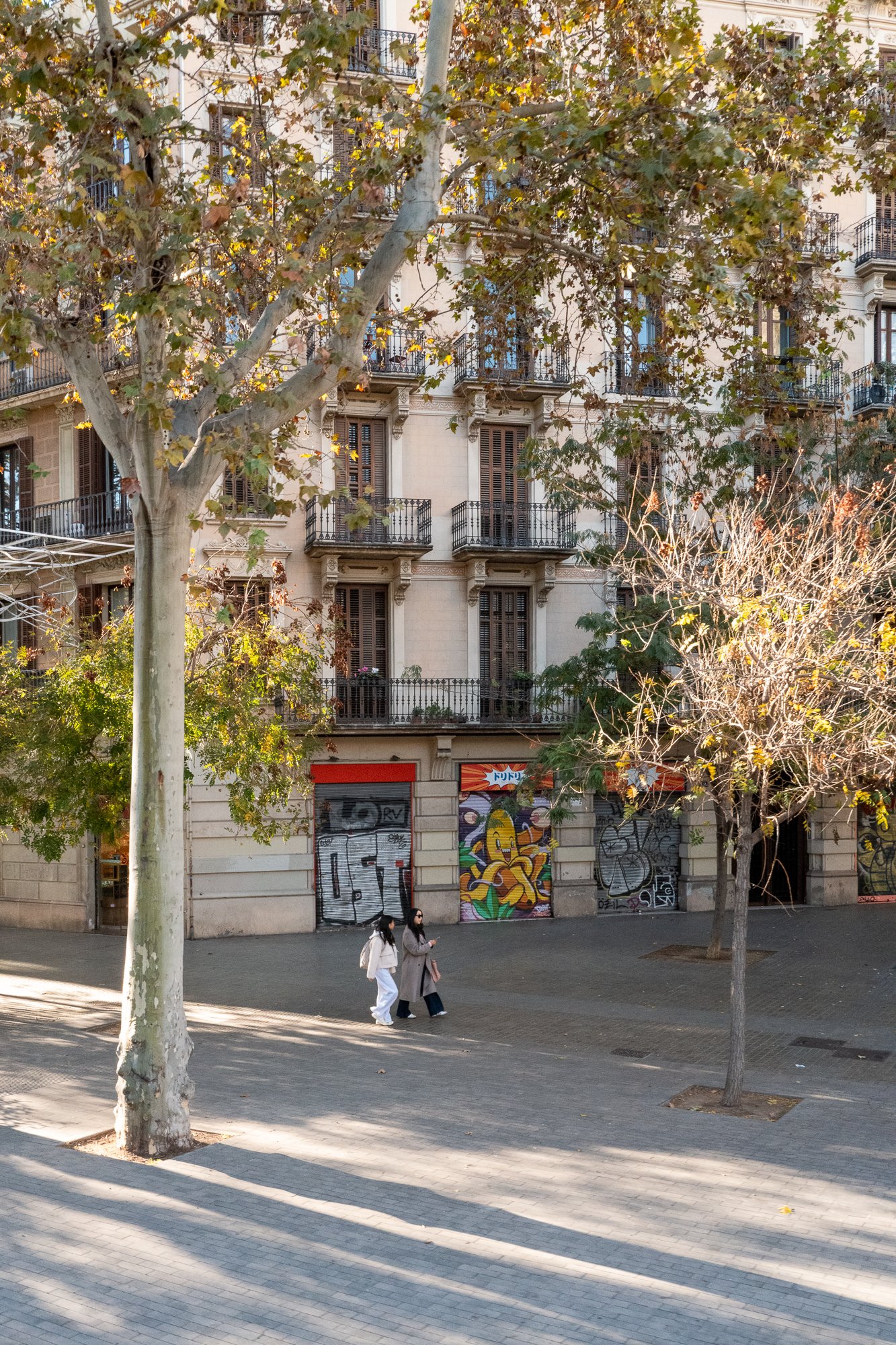 Une rue calme bordée d'arbres avec un immeuble résidentiel à plusieurs étages en arrière-plan, des graffitis colorés sur des volets de magasins fermés au rez-de-chaussée, deux femmes marchant en étant vêtues chaudement, le tout sous une lumière douce