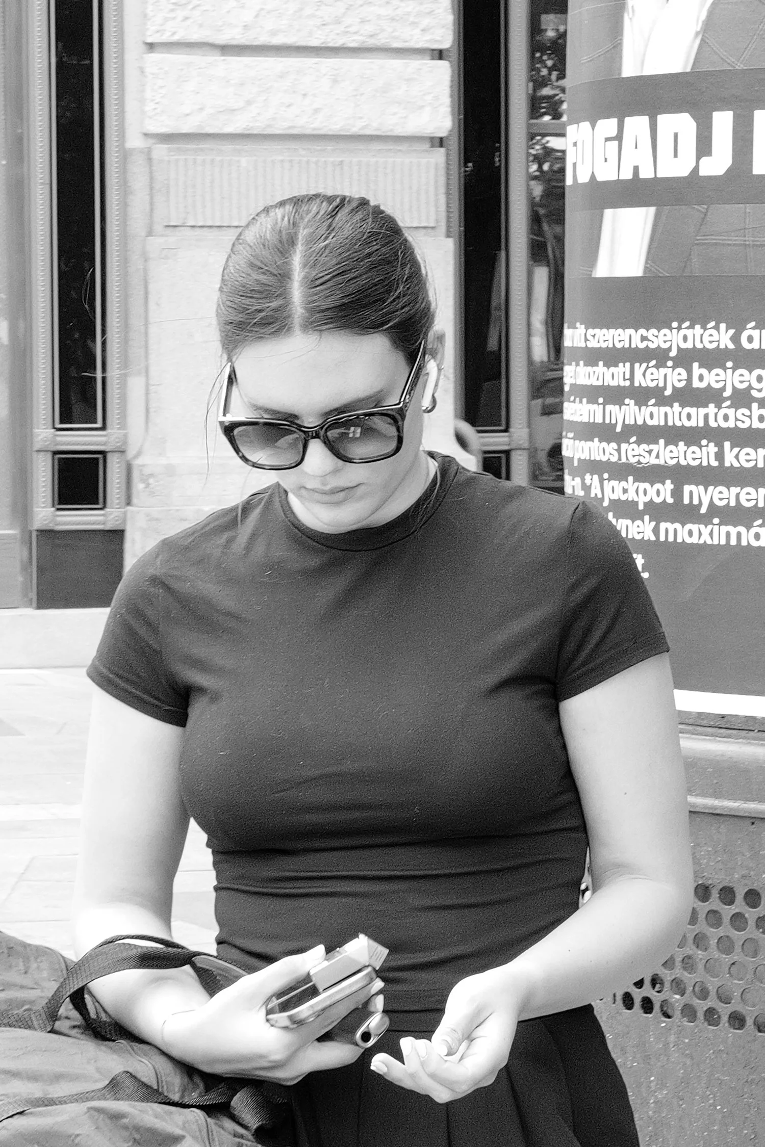 Femme portant des lunettes de soleil, regardant son téléphone, en plein air près d'un bâtiment, en noir et blanc.