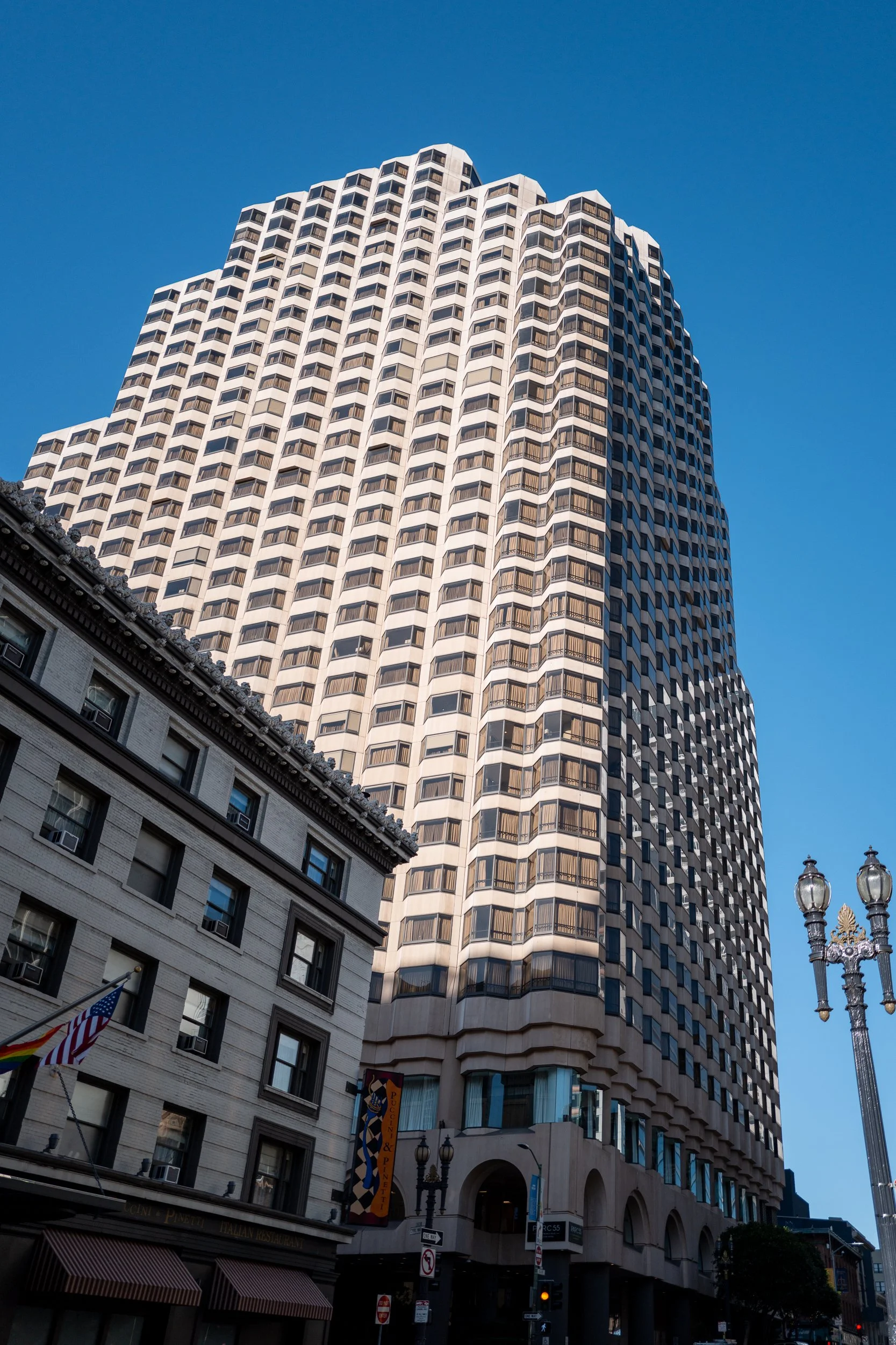 Un grand immeuble moderne en forme de tour avec une façade en forme de grille, contrastant avec un bâtiment plus ancien en pierre au premier plan, sous un ciel bleu clair.