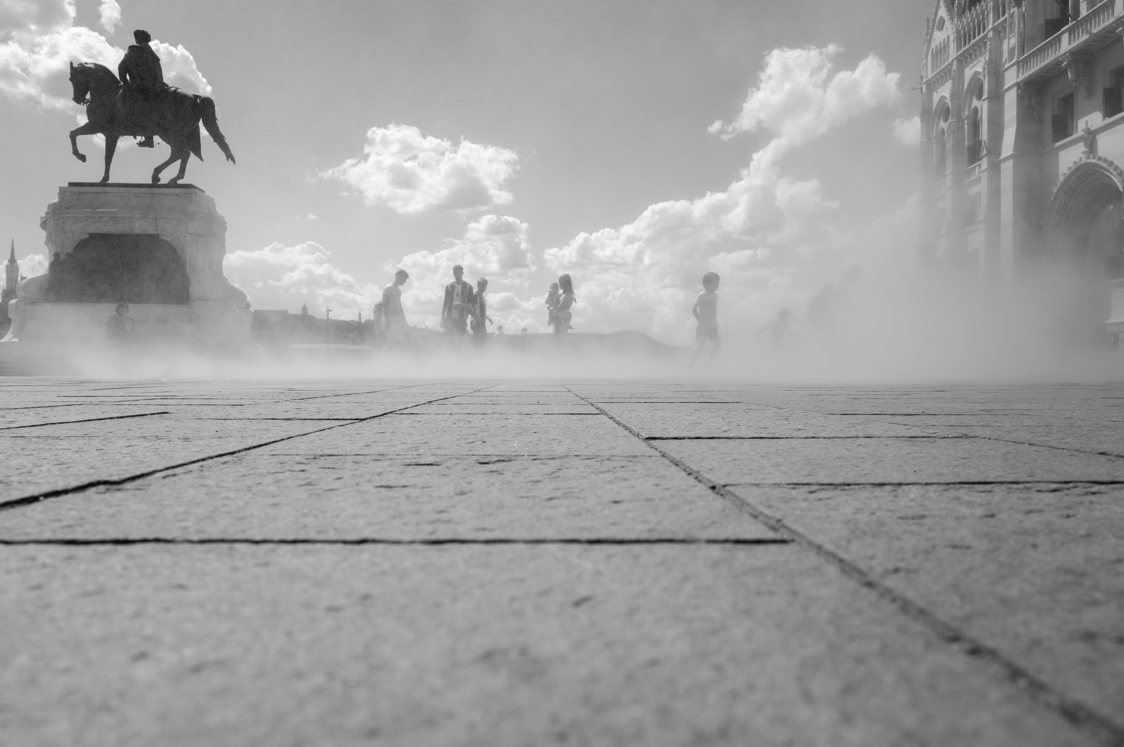 Une place publique avec une statue équestre sur un socle, plusieurs personnes se promènent et un peu de brouillard ou de vapeur monte du sol, avec un bâtiment dans le fond et un ciel partiellement nuageux.