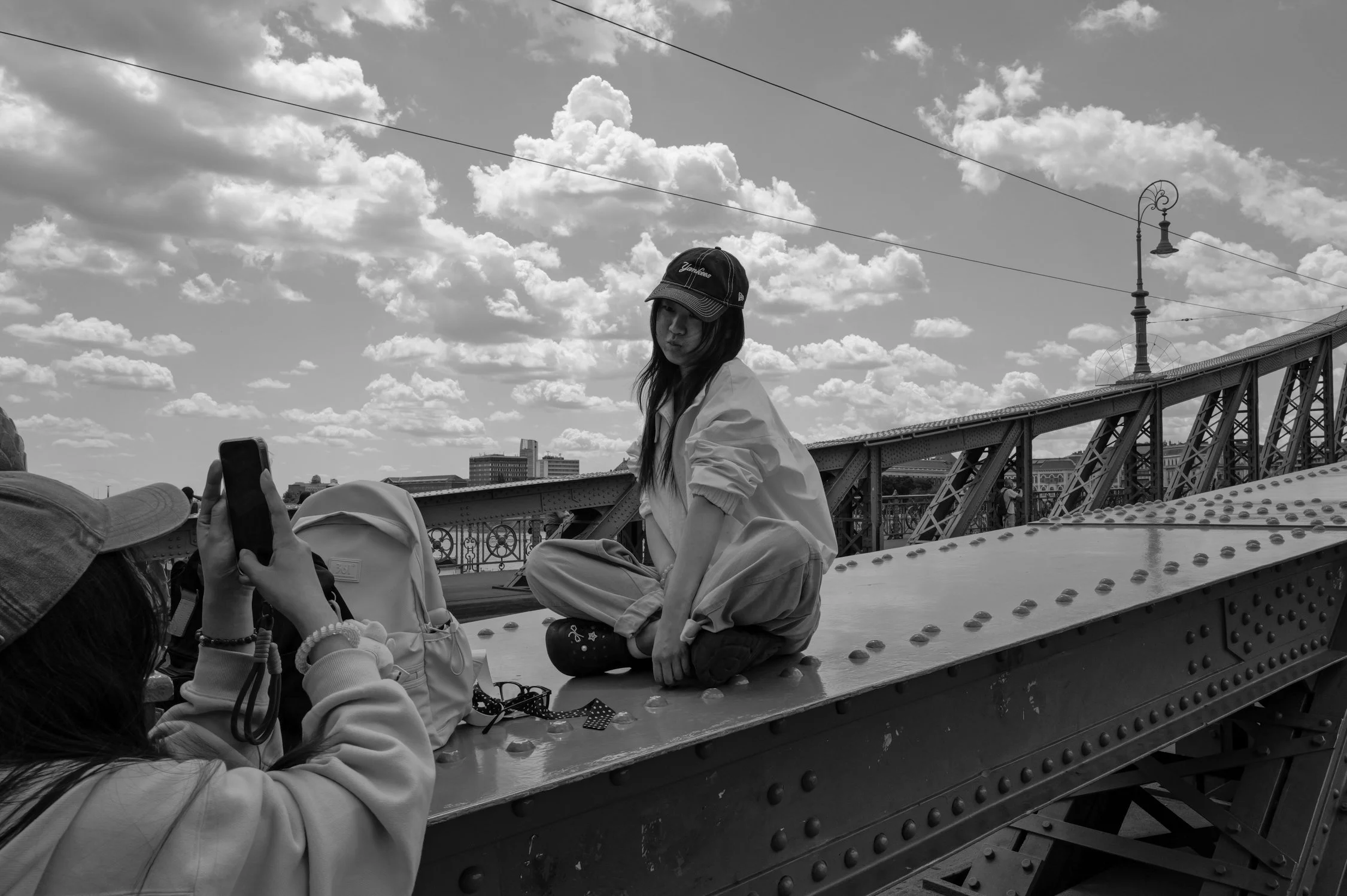 Une jeune femme, portant une casquette et une veste, est assise sur une structure métallique d'un pont, sous un ciel nuageux. Plusieurs personnes prennent des photos ou des selfies.