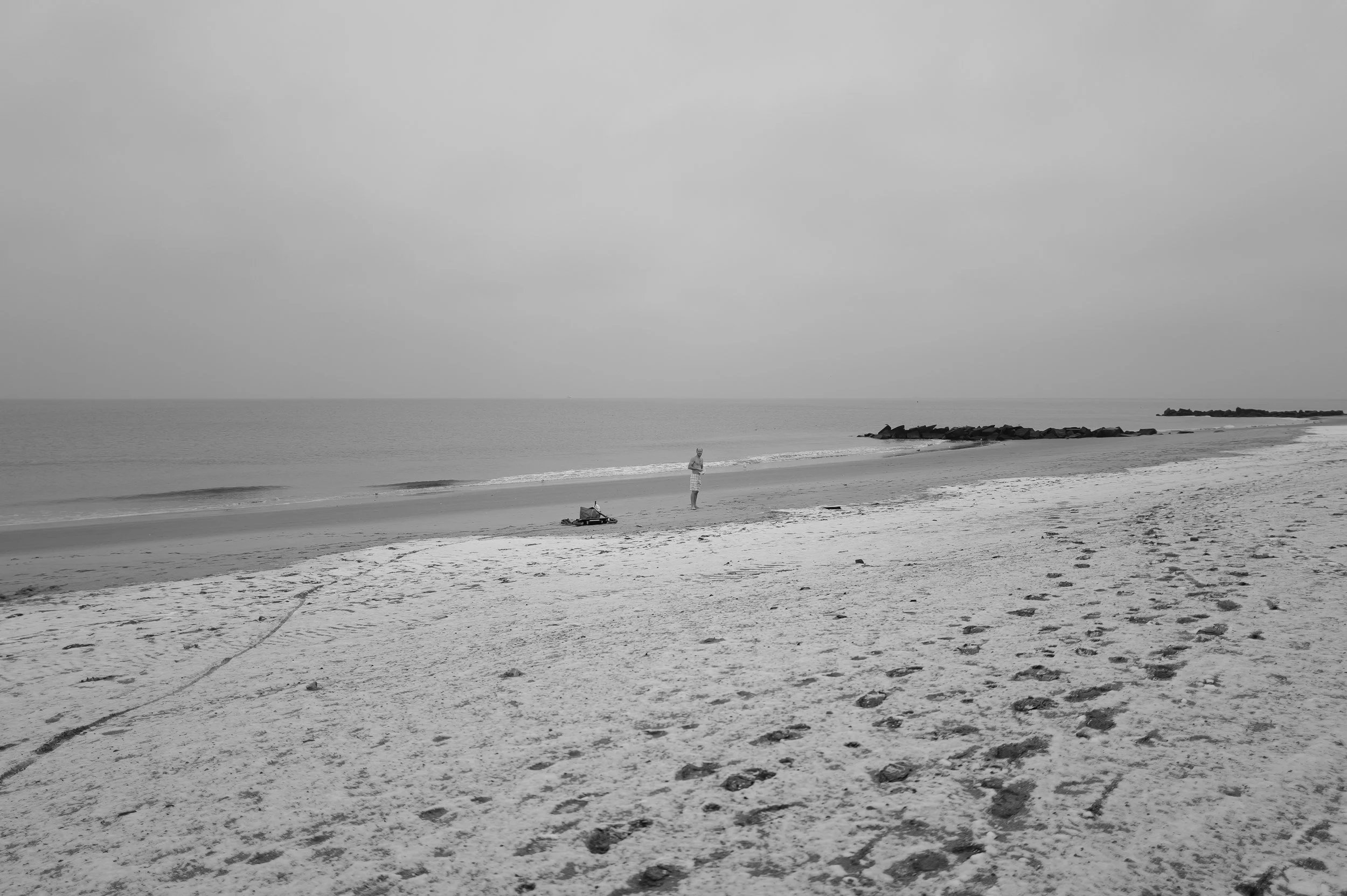 Plage déserte avec un seul personne, quelques animaux en peluche et des bottes sur le sable, avec des rochers au loin, sous un ciel nuageux.