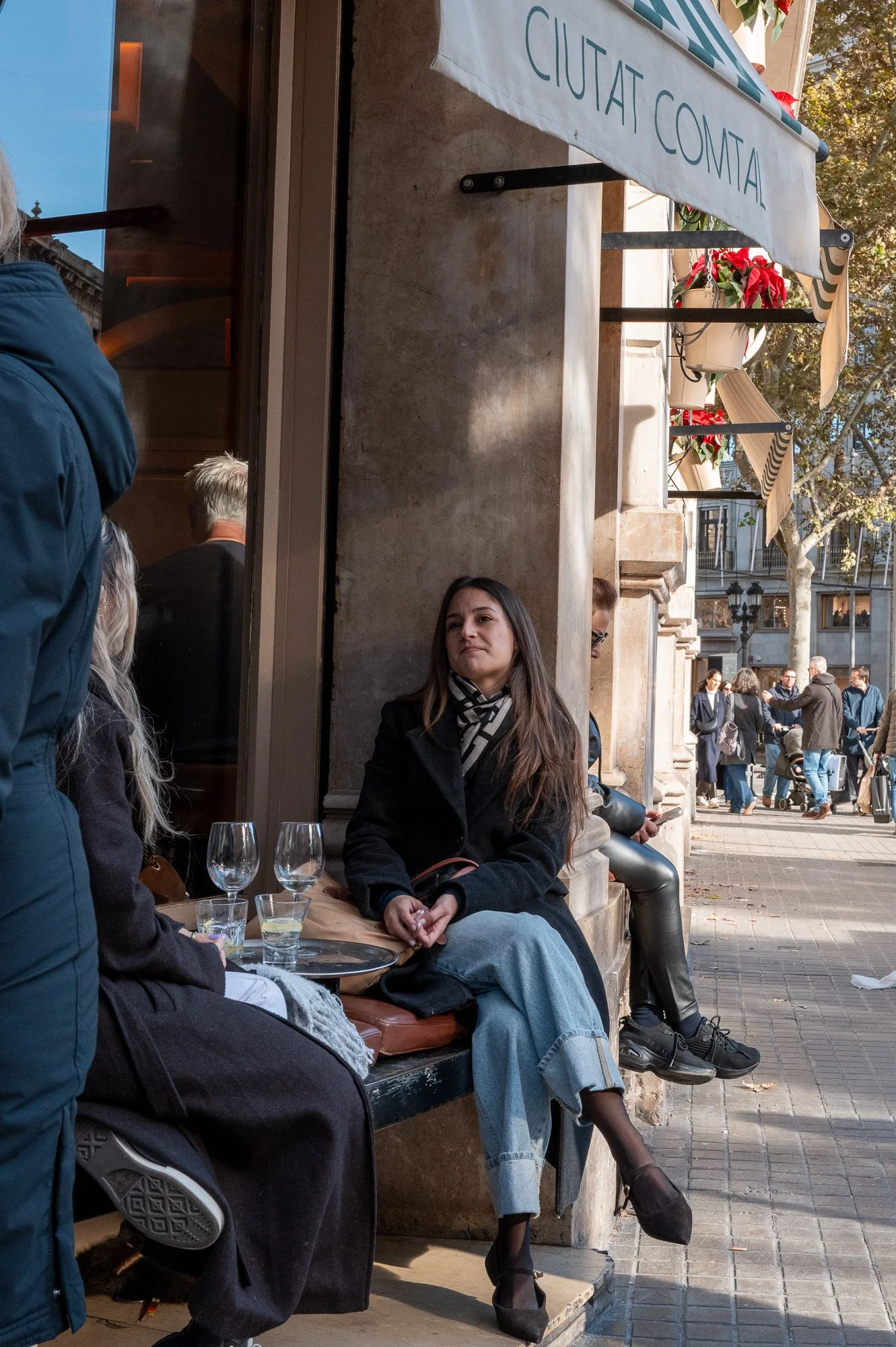 Femme assise sur un banc devant un café, la rue animée en arrière-plan, avec des personnes qui marchent et discutent.