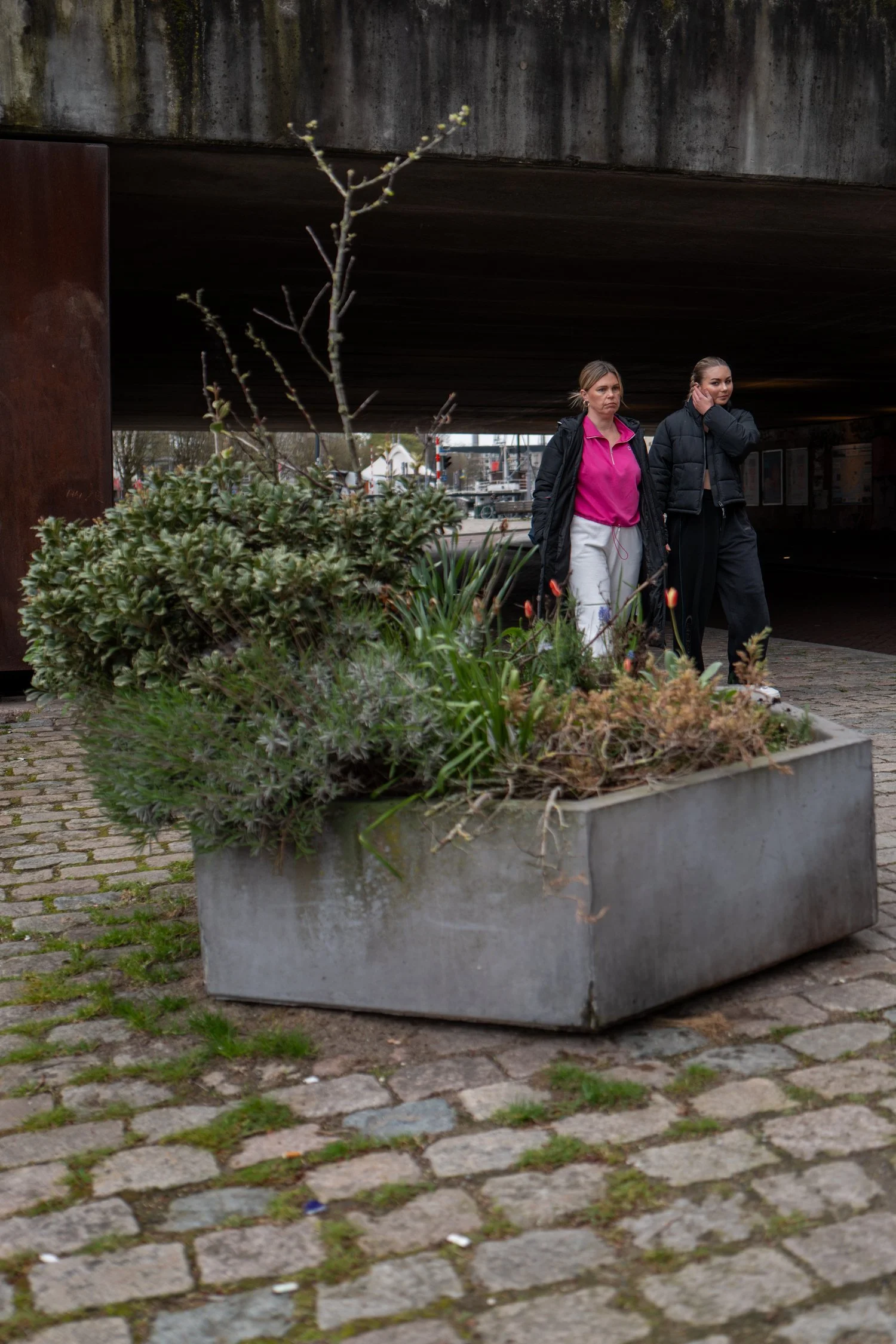 Deux femmes marchent sous un pont en béton, derrière un grand pot de fleurs rempli de végétation.