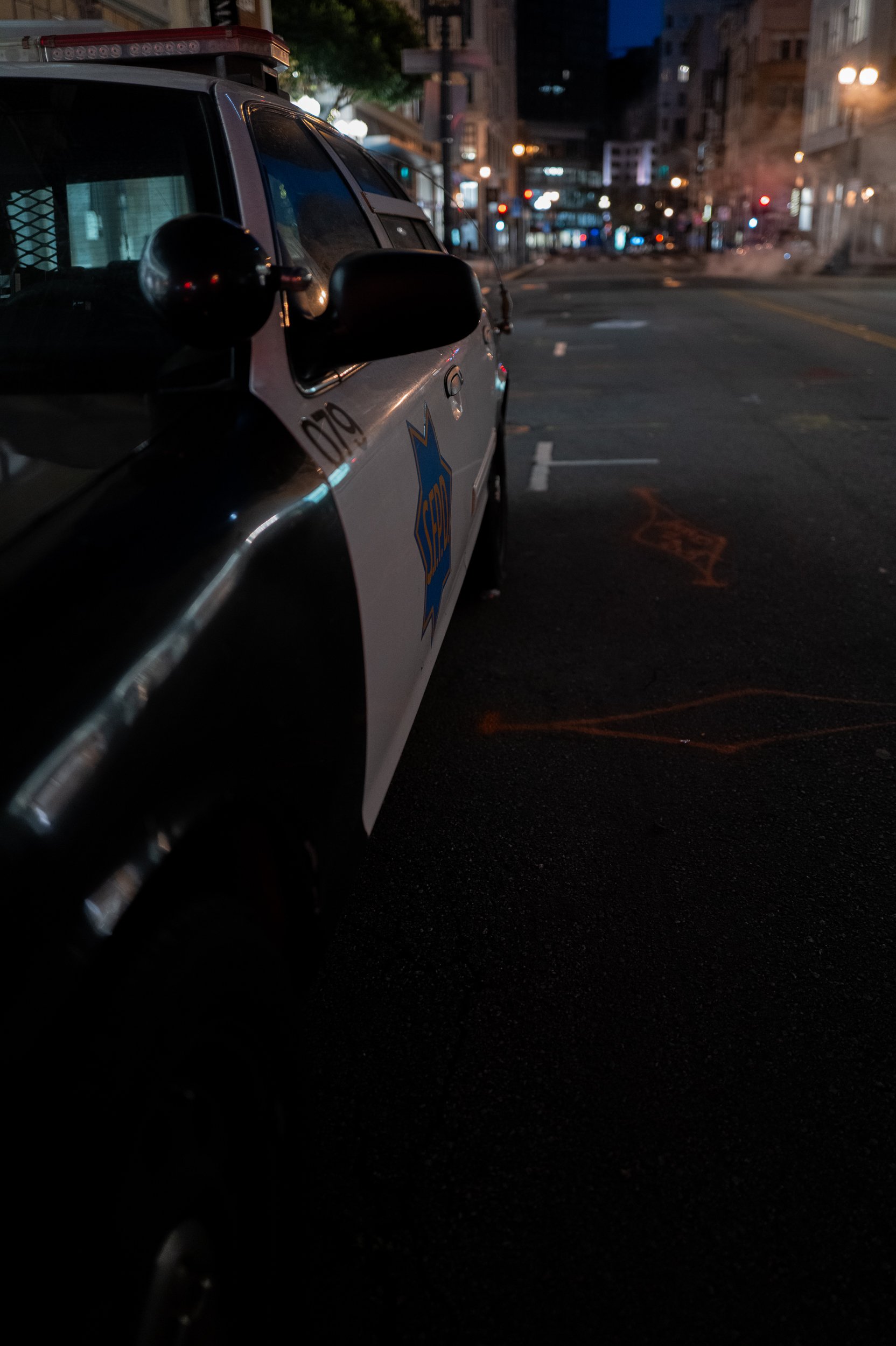 Voiture de police stationnée dans une rue urbaine sombre, avec des bâtiments et des lumières nocturnes en arrière-plan.