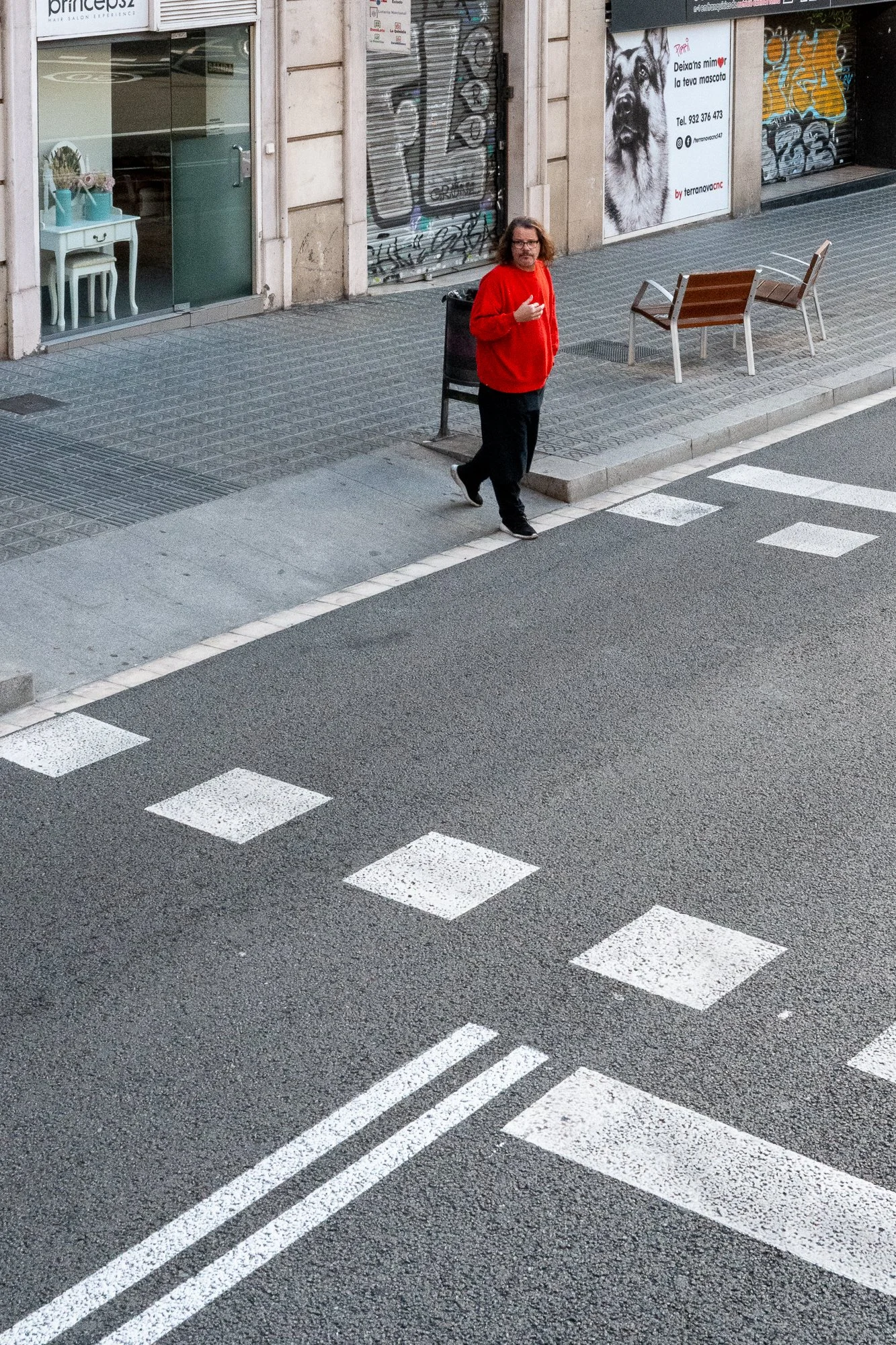 Un homme portant un sweat rouge et des pantalons noirs traverse une rue piétonne. La scène se déroule devant un bâtiment avec une boutique dont la vitrine montre un intérieur avec une petite table blanche et des chaises. Il y a des graffitis sur les 
