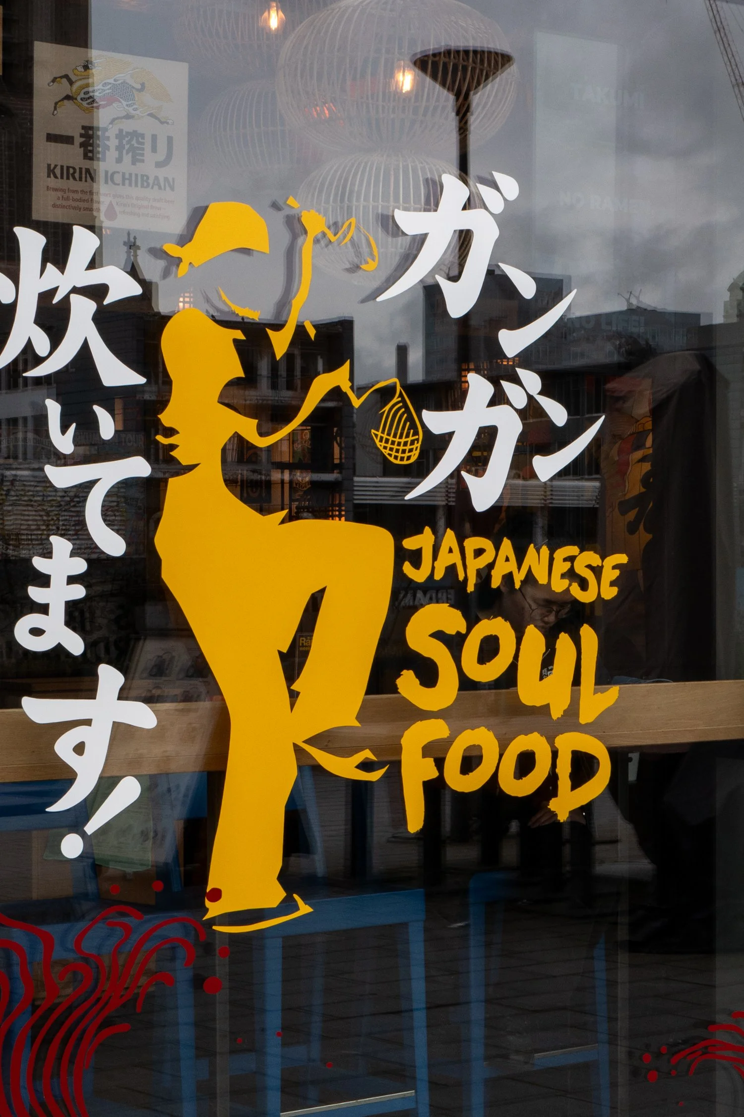 Une fenêtre de restaurant avec une illustration d'une femme en jaune, d'une bouteille de saké, et du texte en anglais et en japonais annonçant la nourriture japonaise.