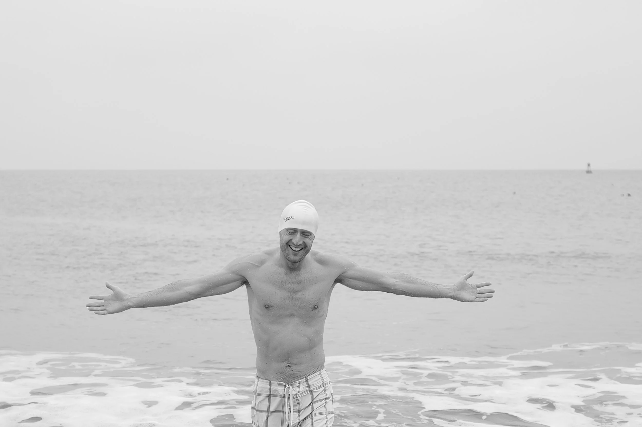 Un homme nageant dans la mer, portant un bonnet de bain, souriant avec les bras ouverts, avec l'océan en arrière-plan.