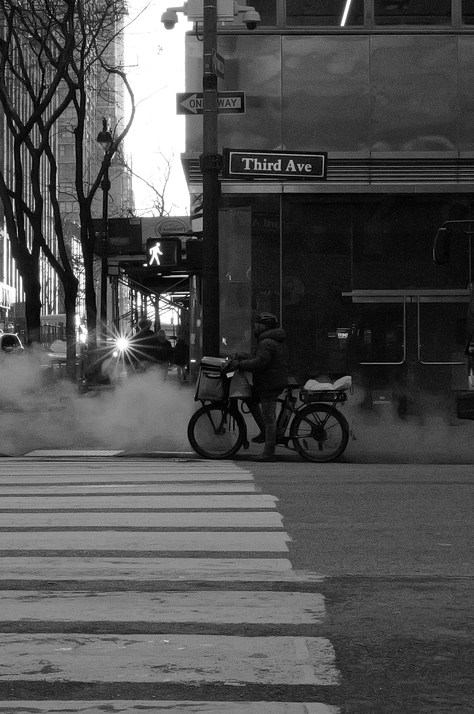 Un homme à vélo attend au passage pour piétons sous un feu piéton, avec une affiche de rue indiquant « Third Ave » et un panneau « One Way » visible en arrière-plan. Il fait froid, de la vapeur ou de la fumée s'élève du sol, et il y a des arbres et d
