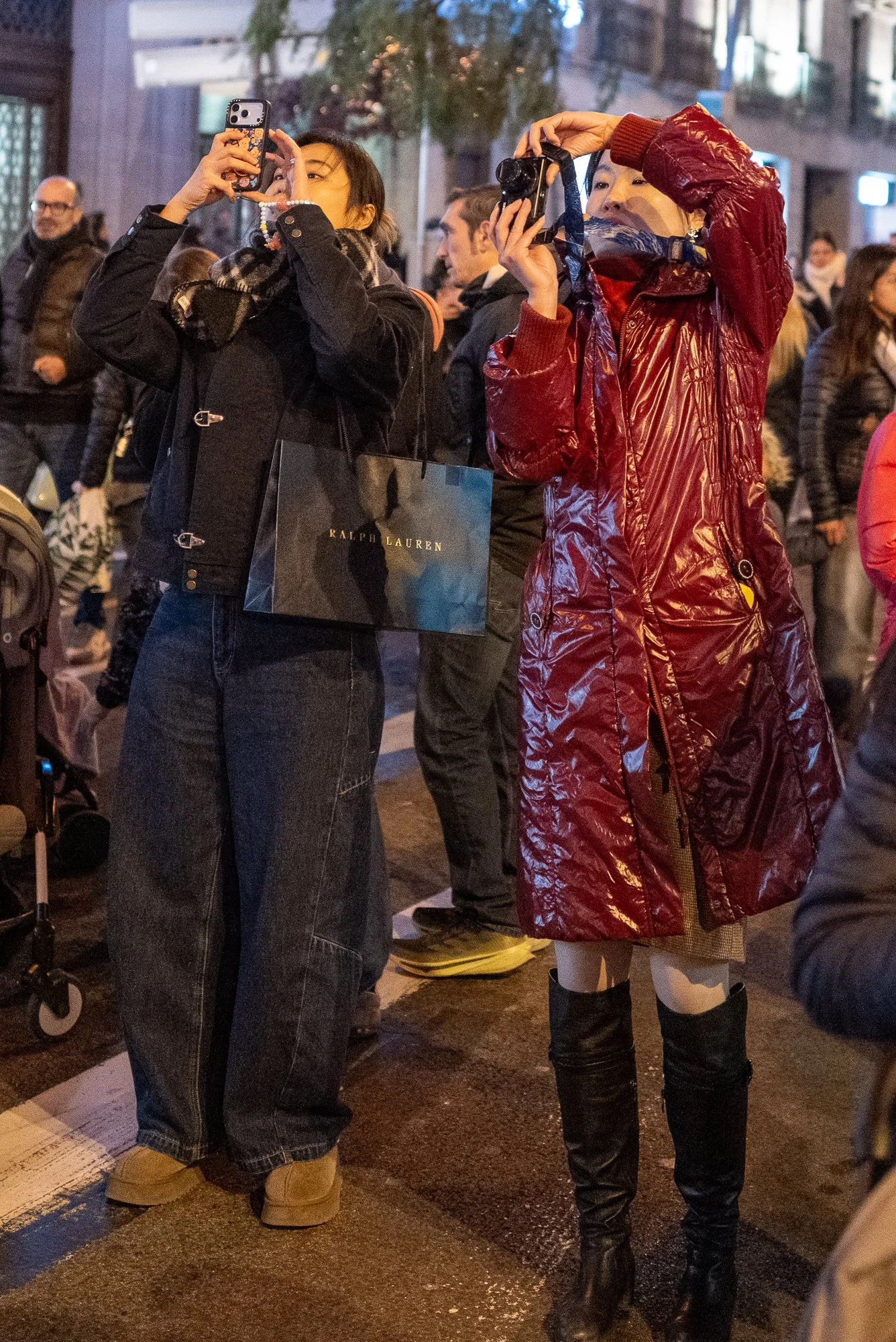 Deux femmes prennent des photos avec leur téléphone dans une rue bondée la nuit, entourées d'autres personnes. L'une porte un manteau noir avec un sac Ralph Lauren, l'autre une veste rouge en plastique avec des bottes hautes noires.