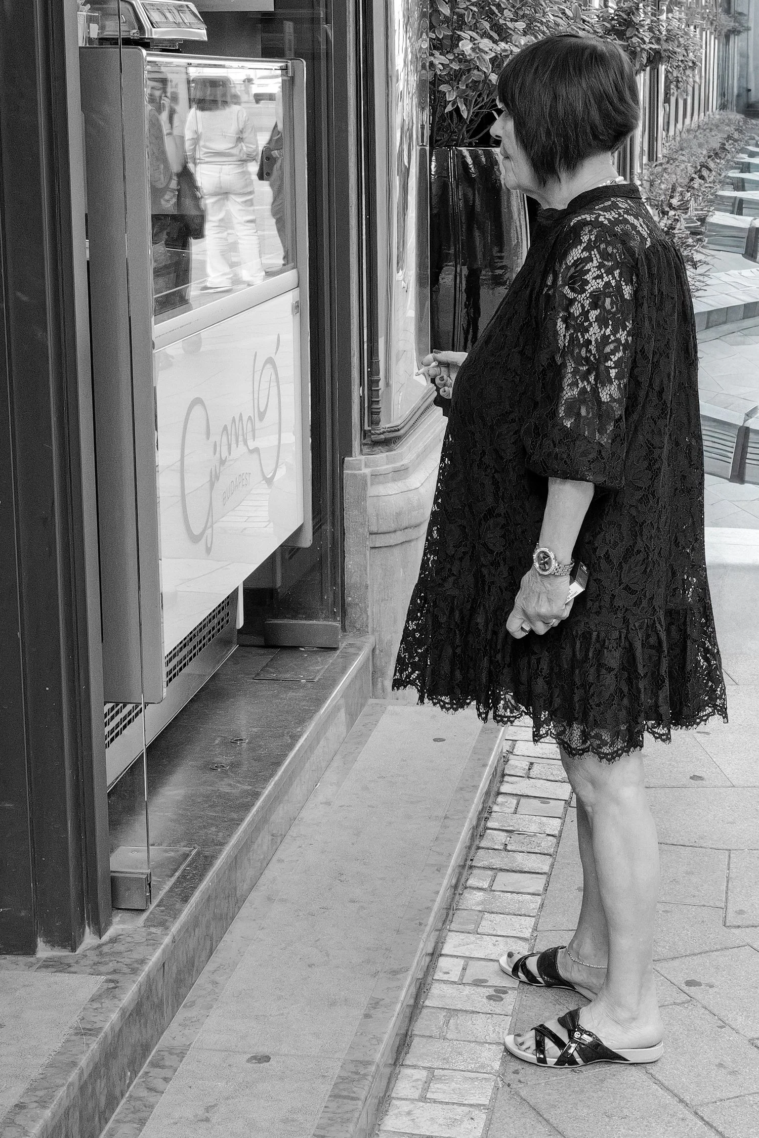 Femme âgée de dos, habillée d'une robe en dentelle noire, se tenant à une machine à billets dans une rue pavée.