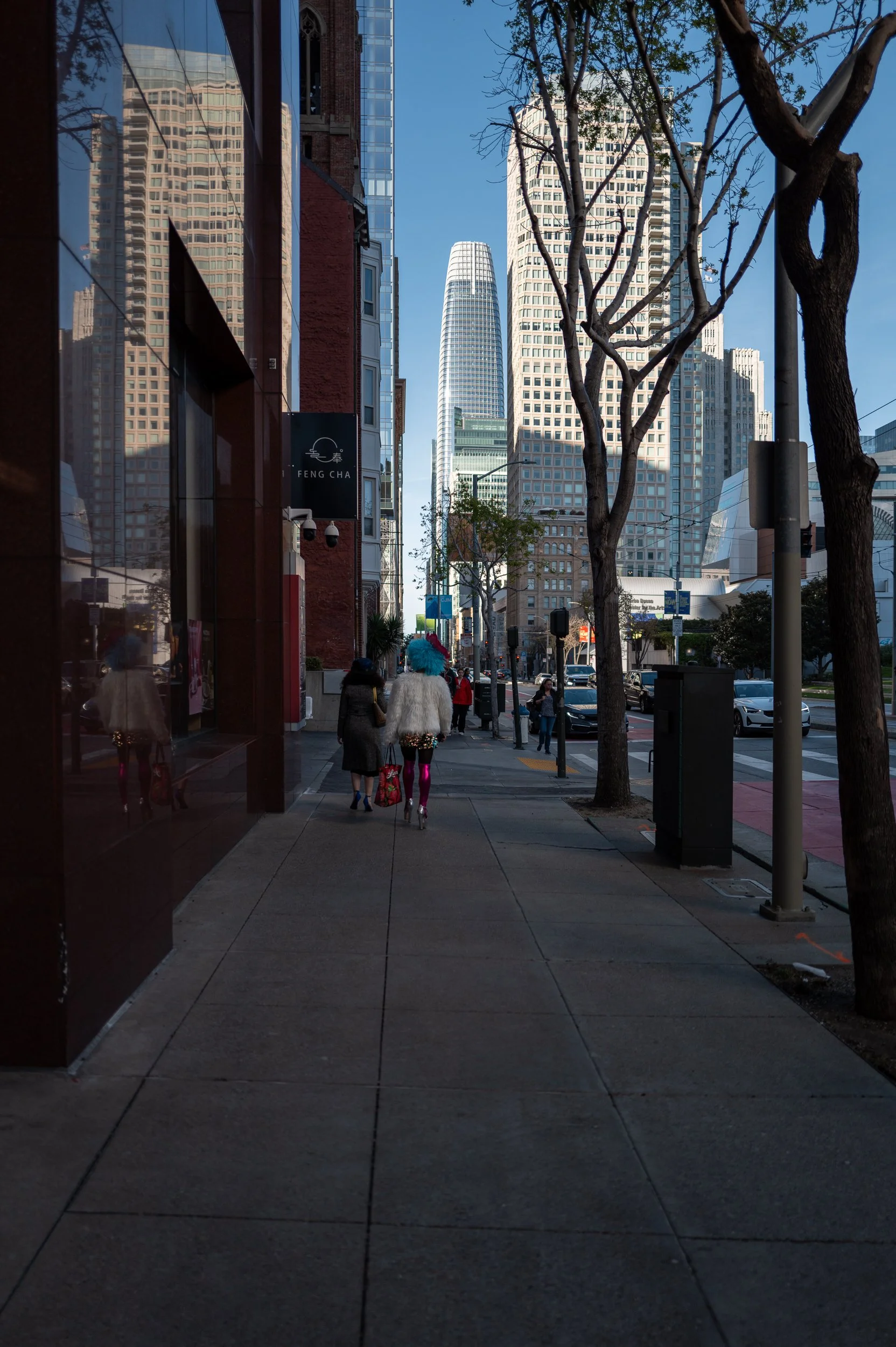 Une rue urbaine avec des gratte-ciel modernes, des arbres et des piétons, dont une personne en costume extravagant avec une perruque bleue et des collants roses, marchant côte à côte.