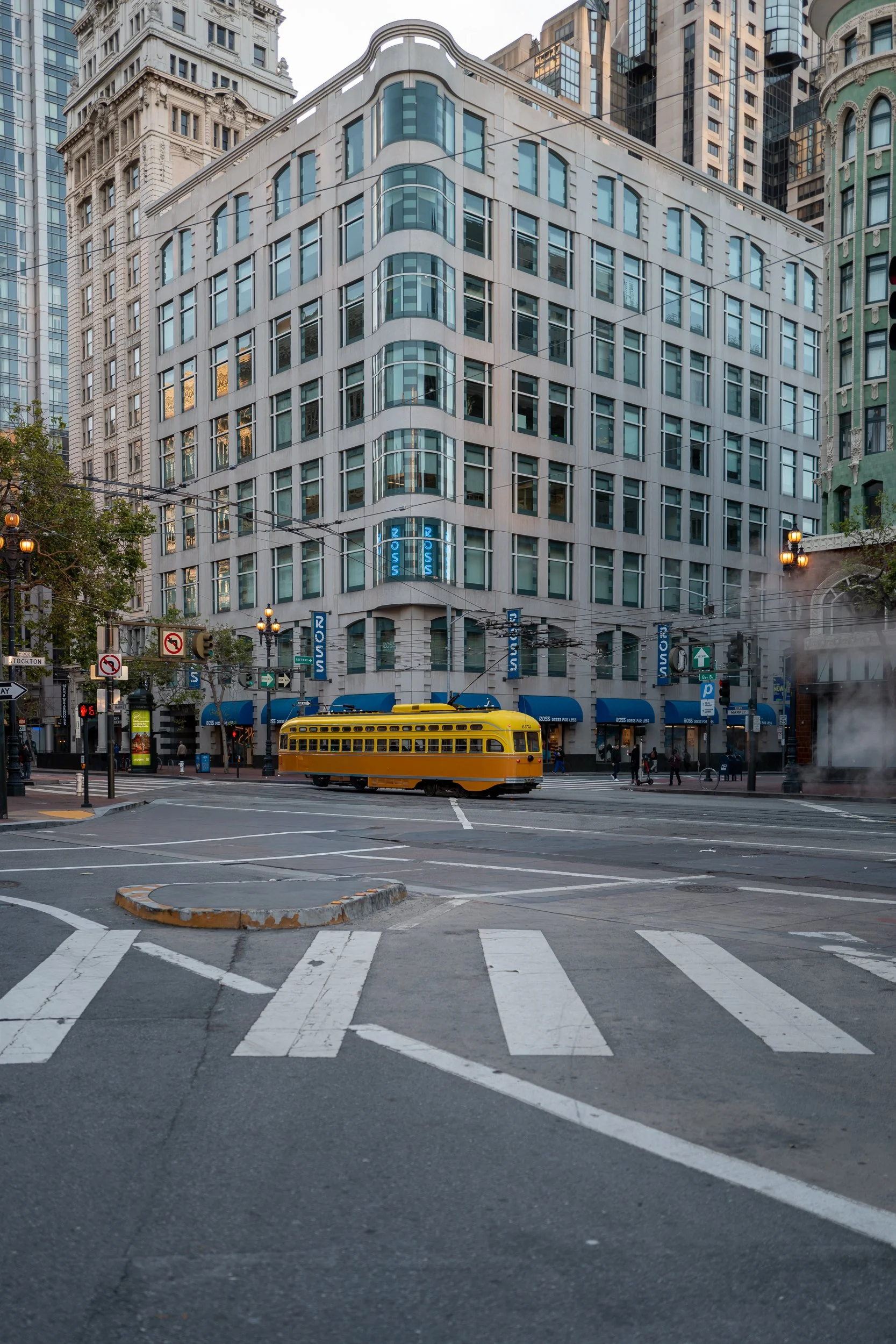 Une rue urbaine avec un tramway jaune passant devant un immeuble moderne en verre et en pierre, avec des rues piétonnes et des lampadaires en arrière-plan.