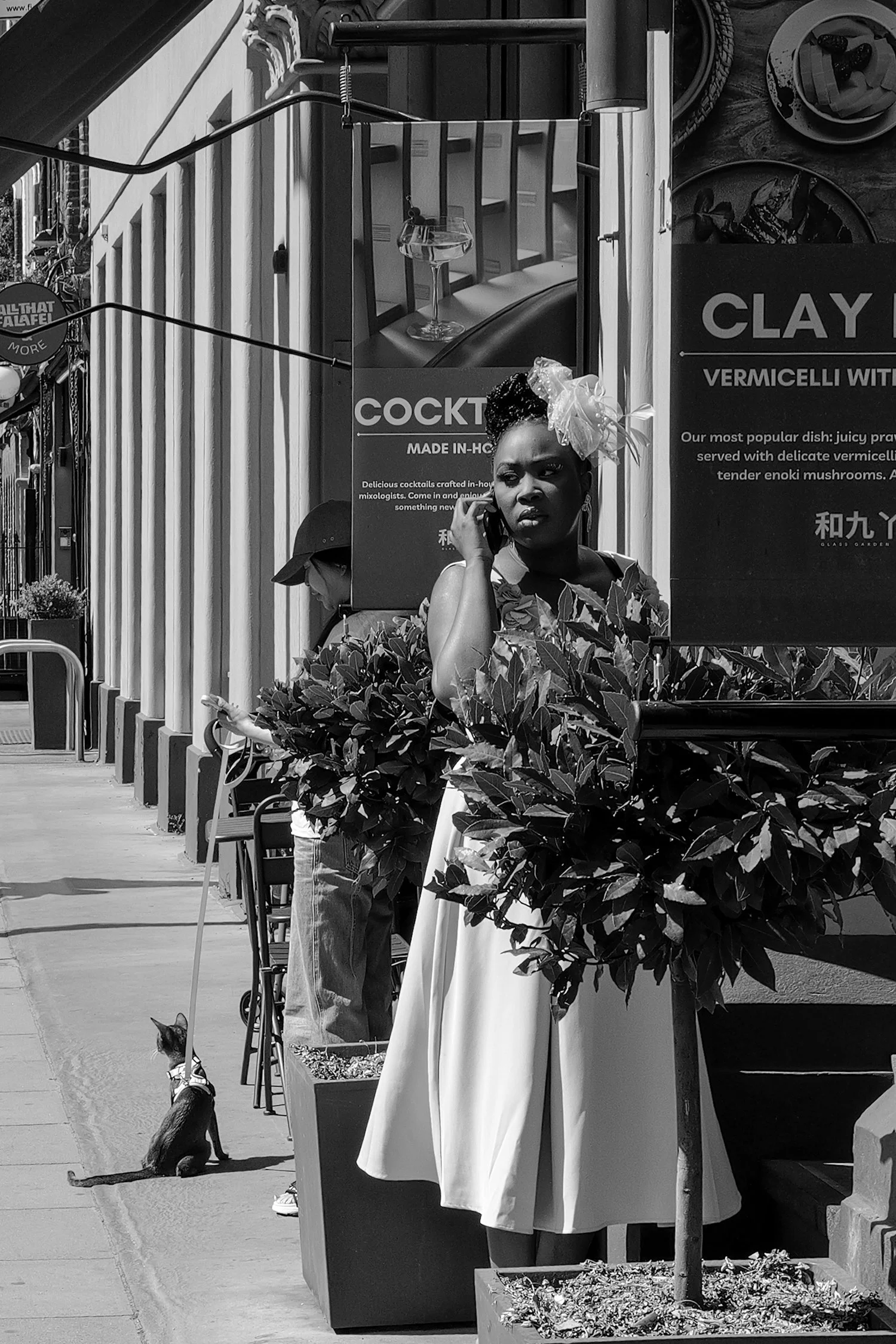 Une femme portant une coiffure décorative et une robe blanche, tenant un téléphone, se trouve sous un parasol près d'une jardinière avec des plantes, dans une zone urbaine avec des affiches publicitaires en arrière-plan.