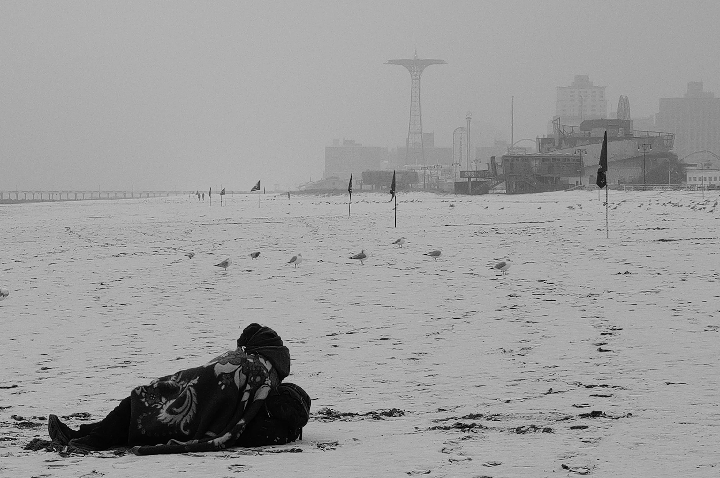 Une personne assise sur la plage enneigée, portant une couverture. Le ciel est gris et brumeux, avec des oiseaux sur le sable, et des bâtiments industriels en arrière-plan.