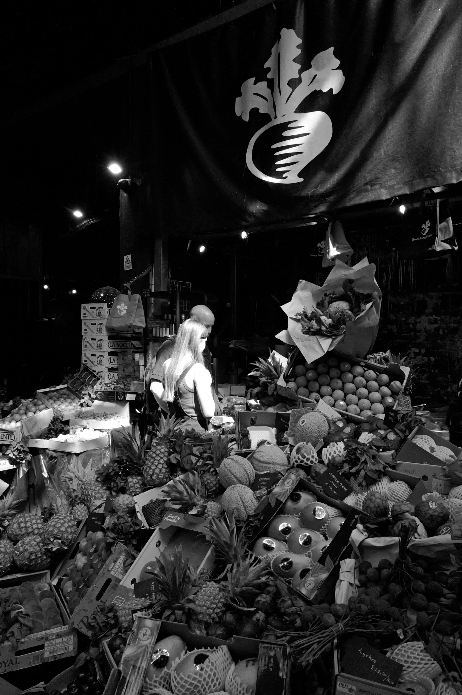 Marché de fruits avec paniers de fruits, notamment ananas, melons, et autres fruits tropicaux, sous un barnum avec logo de carotte.