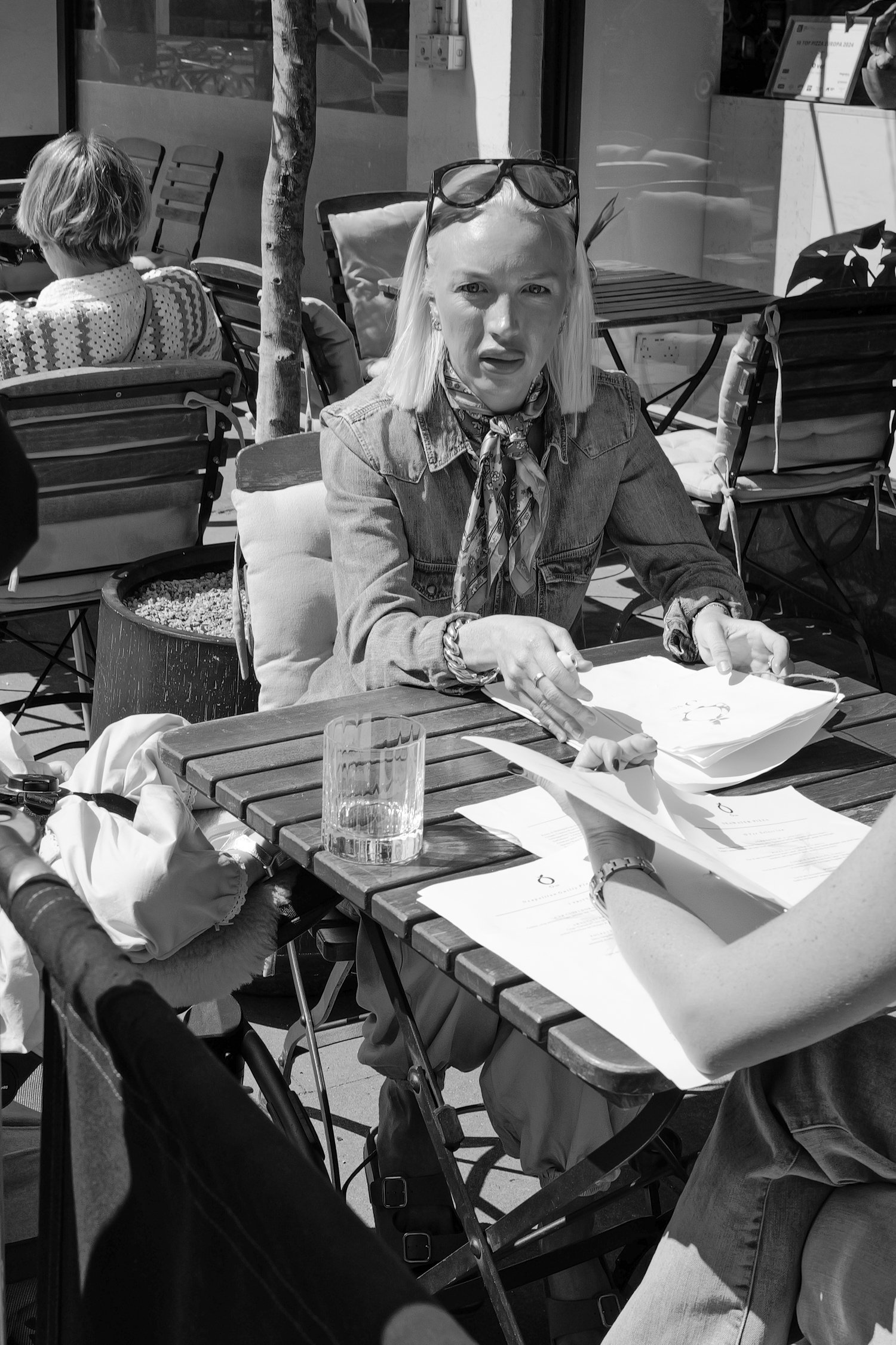 Une femme blonde portant des lunettes de soleil sur la tête, assise à une table en extérieur, avec une paire de lunettes de soleil sur sa tête, en train de feuilleter un menu ou un document, avec un verre d'eau devant elle. Autour, d'autres personnes
