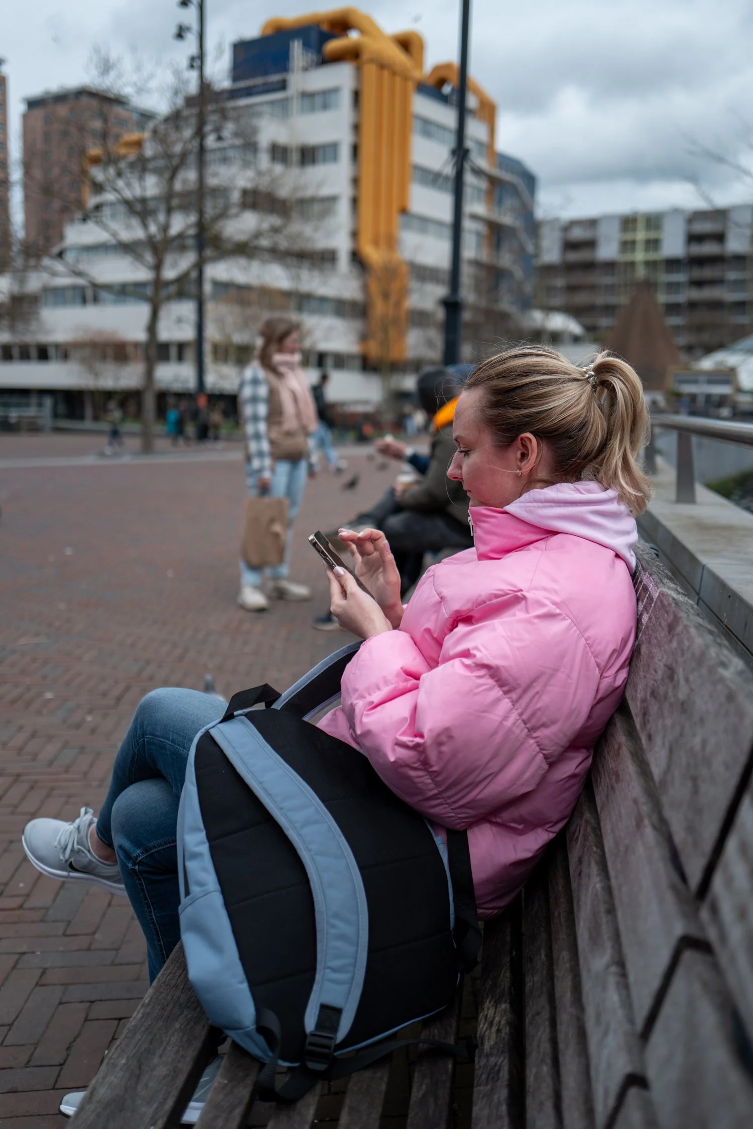 Une femme en manteau rose assise sur un banc, utilisant son téléphone, avec un sac à dos posé sur ses jambes, dans un espace public urbain avec des bâtiments modernes et d'autres personnes en arrière-plan.