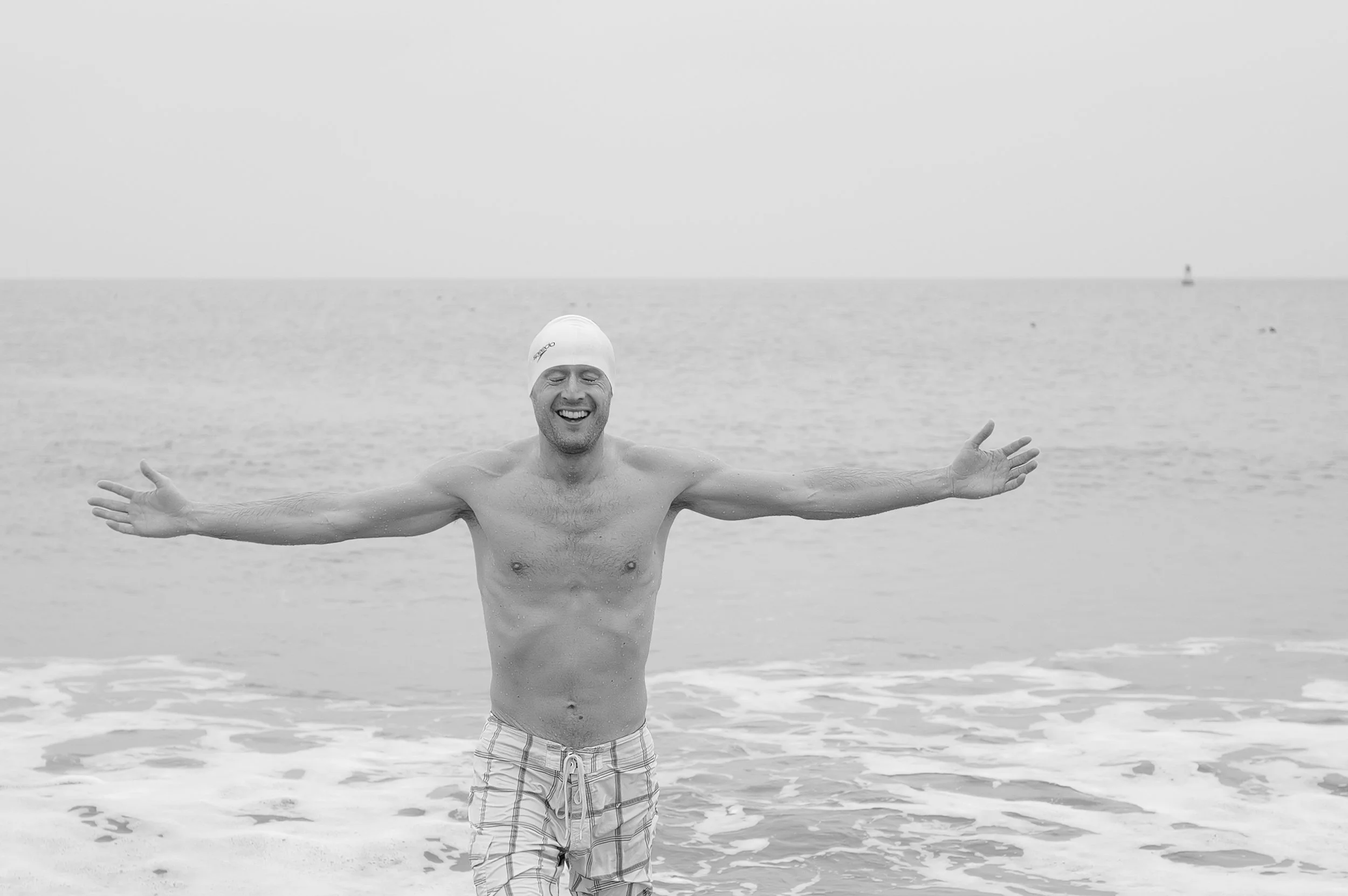 Un homme souriant portant un bonnet de bain, debout dans la mer avec les bras ouverts.