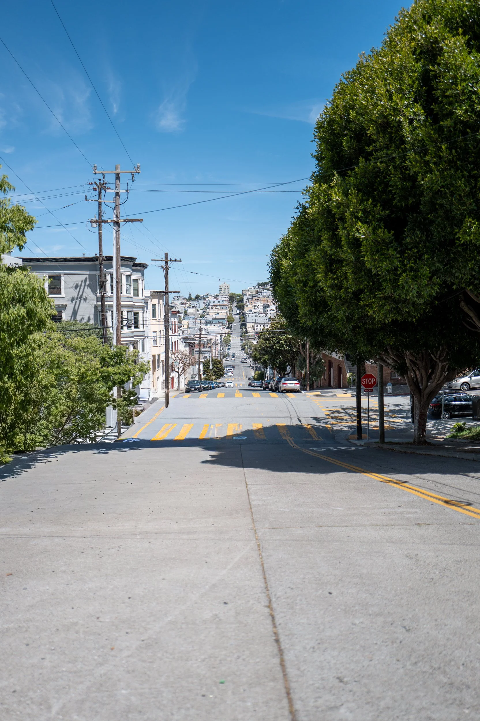 Une rue en pente dans une ville avec des maisons colorées, des arbres, un poteau électrique, un panneau stop, et le ciel bleu.
